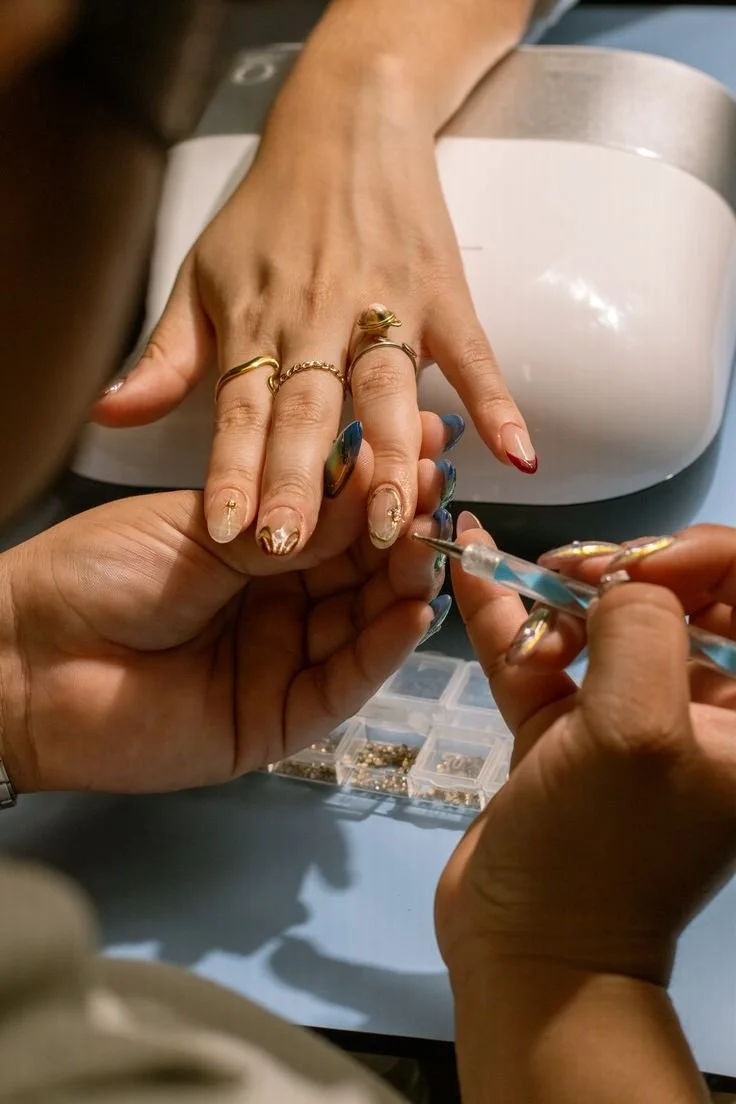 A person receiving a manicure, with their nails being decorated with gold accents, while another person applies clear nail polish with a small brush. There are jewelry pieces on the person's fingers and small containers of nail decor in the background.