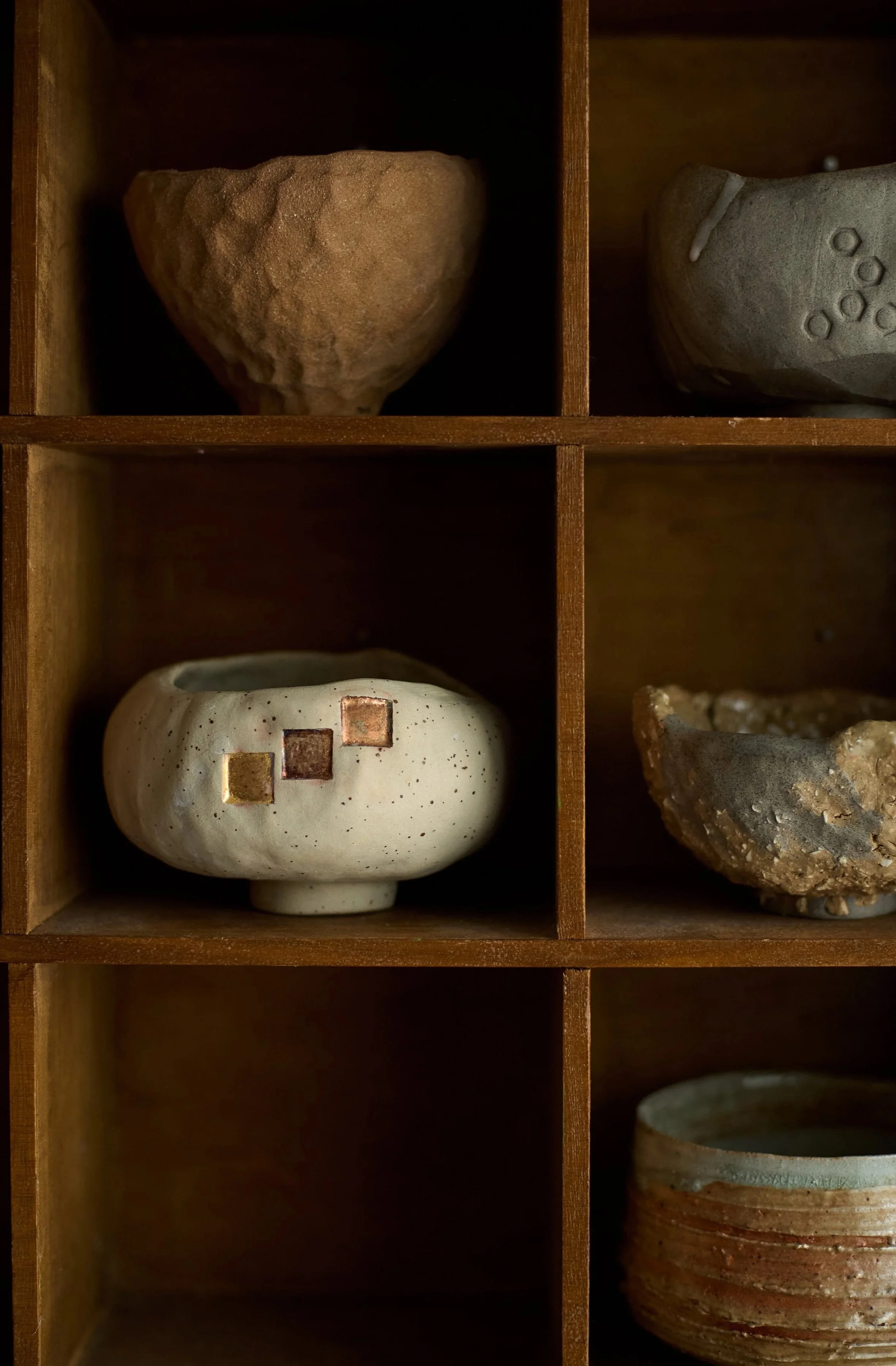 A wooden shelf with six cubbyholes, each containing a small ceramic or stone bowl or vessel of various shapes and textures.