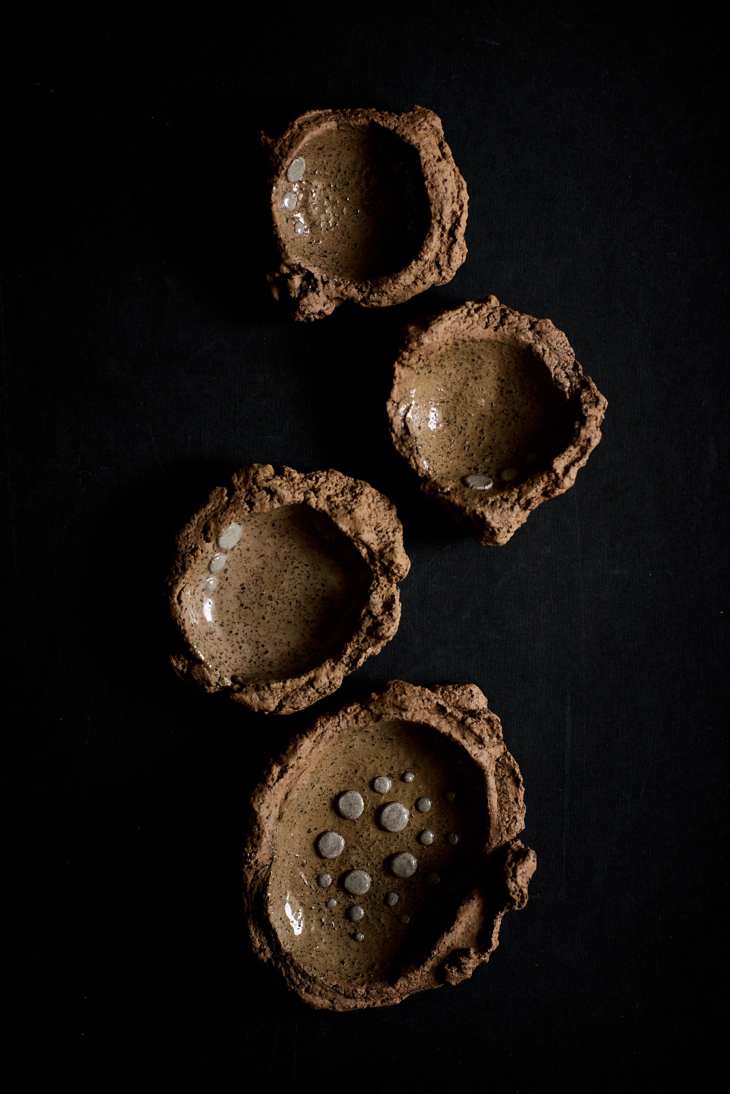 Four small square clay bowls with rough edges and bubbles inside, arranged diagonally on a black surface.