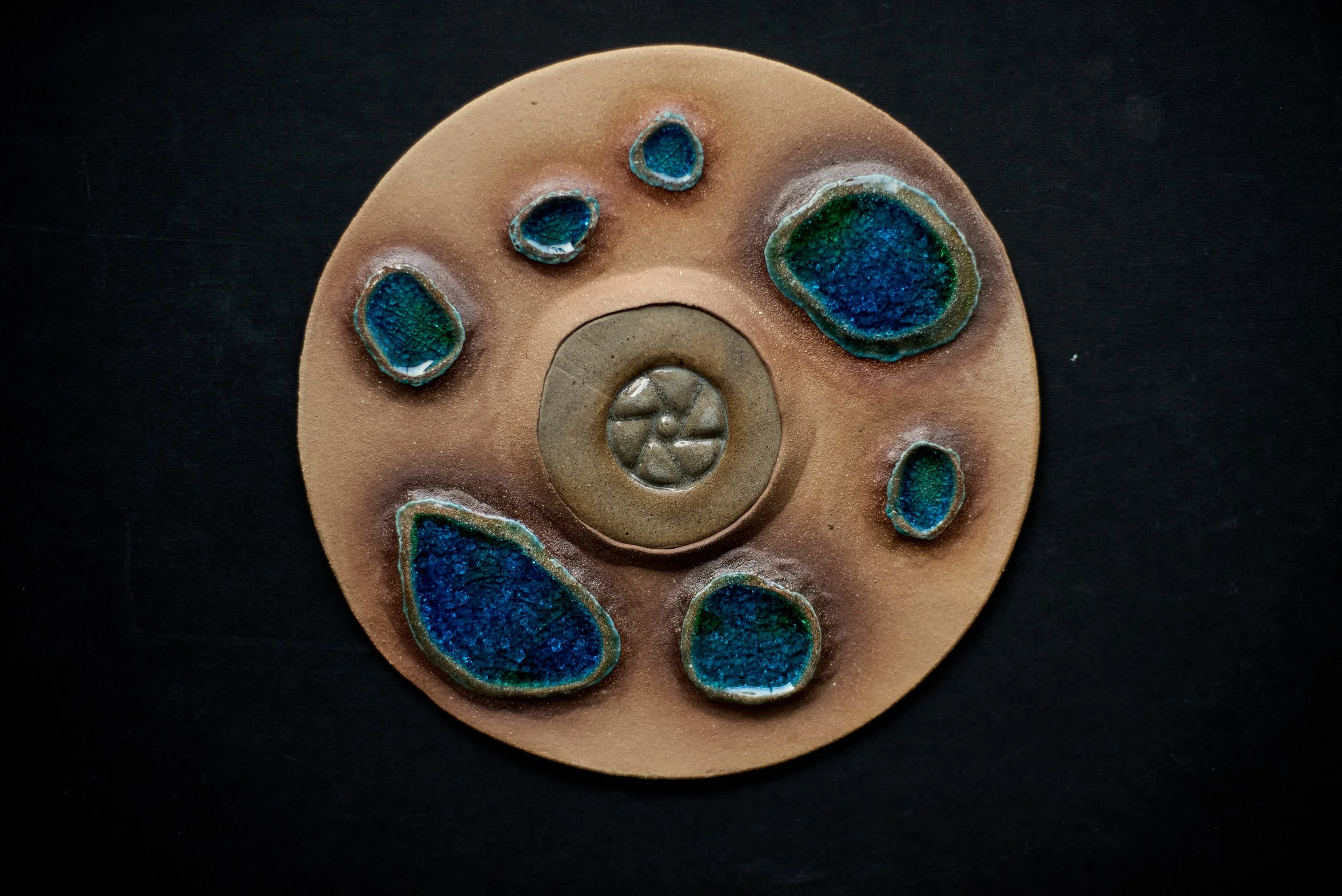A top view of a ceramic or decorative item with several embedded blue and green crystalline or textured stones, set against a dark background.