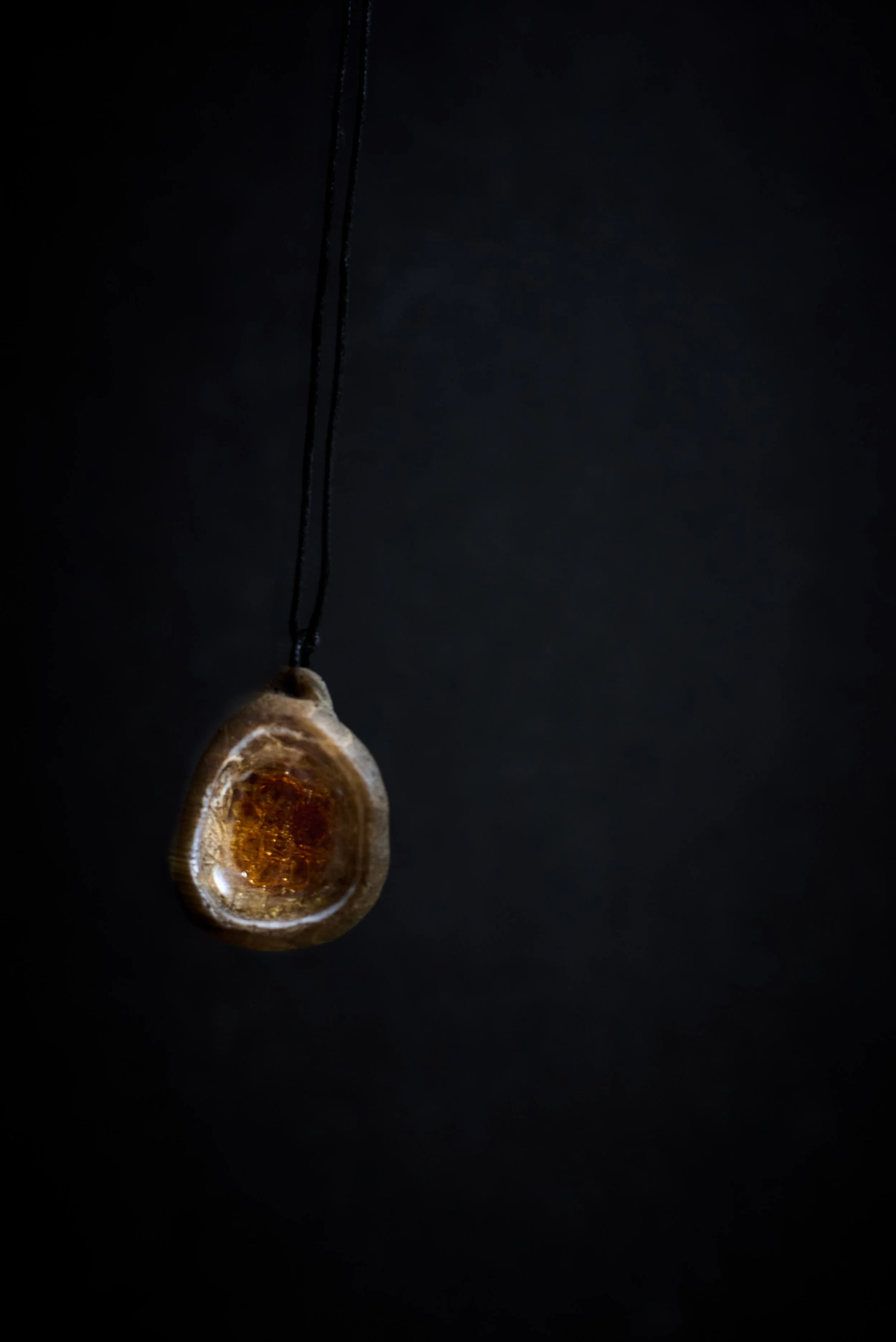 A pendant necklace with a polished amber stone on a black cord, hanging against a dark background.