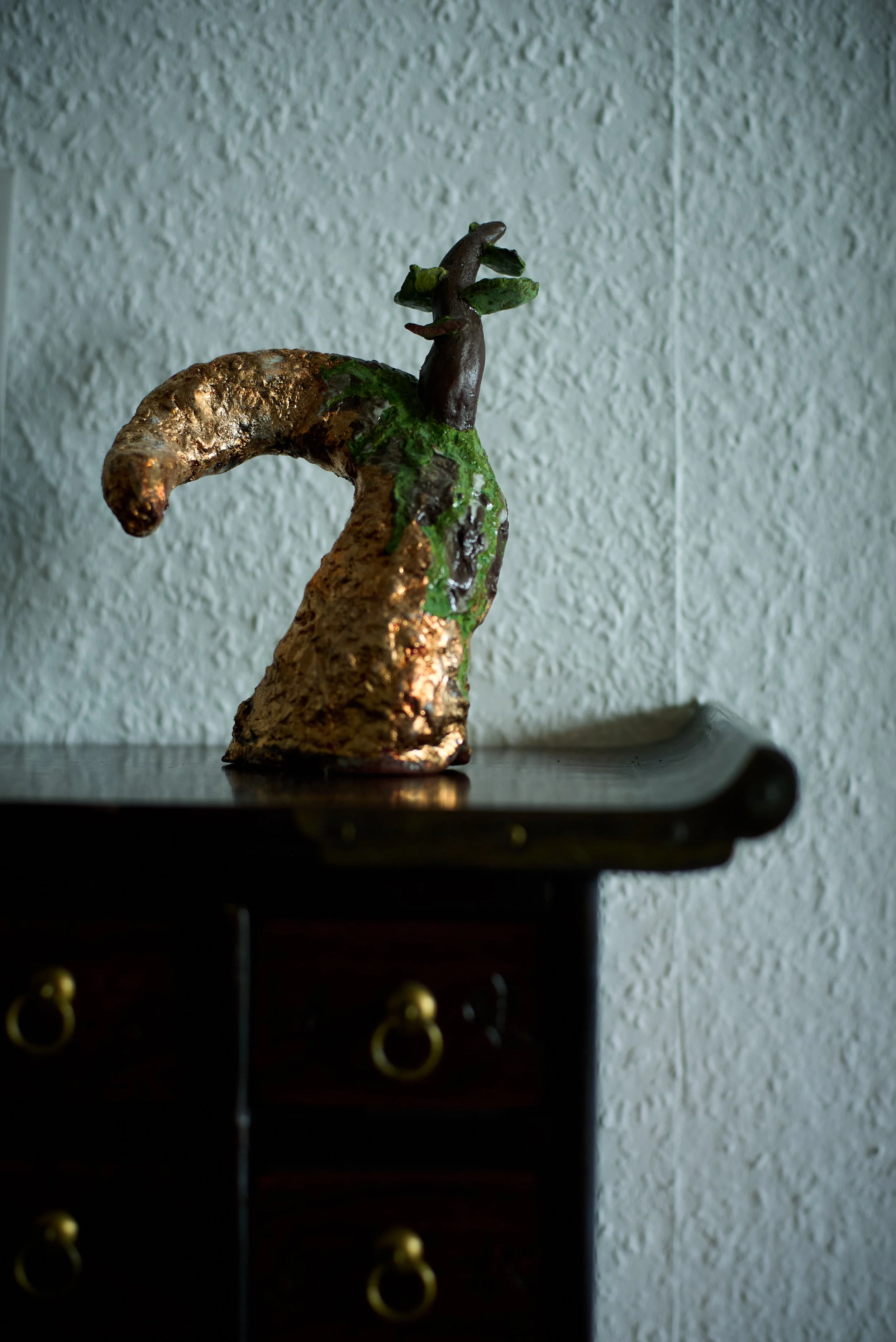 Unique sculpture of a whimsical creature with a brown, curved body and dark brown head featuring protruding branches or horns, displayed on a dark wooden surface against a textured off-white wall.