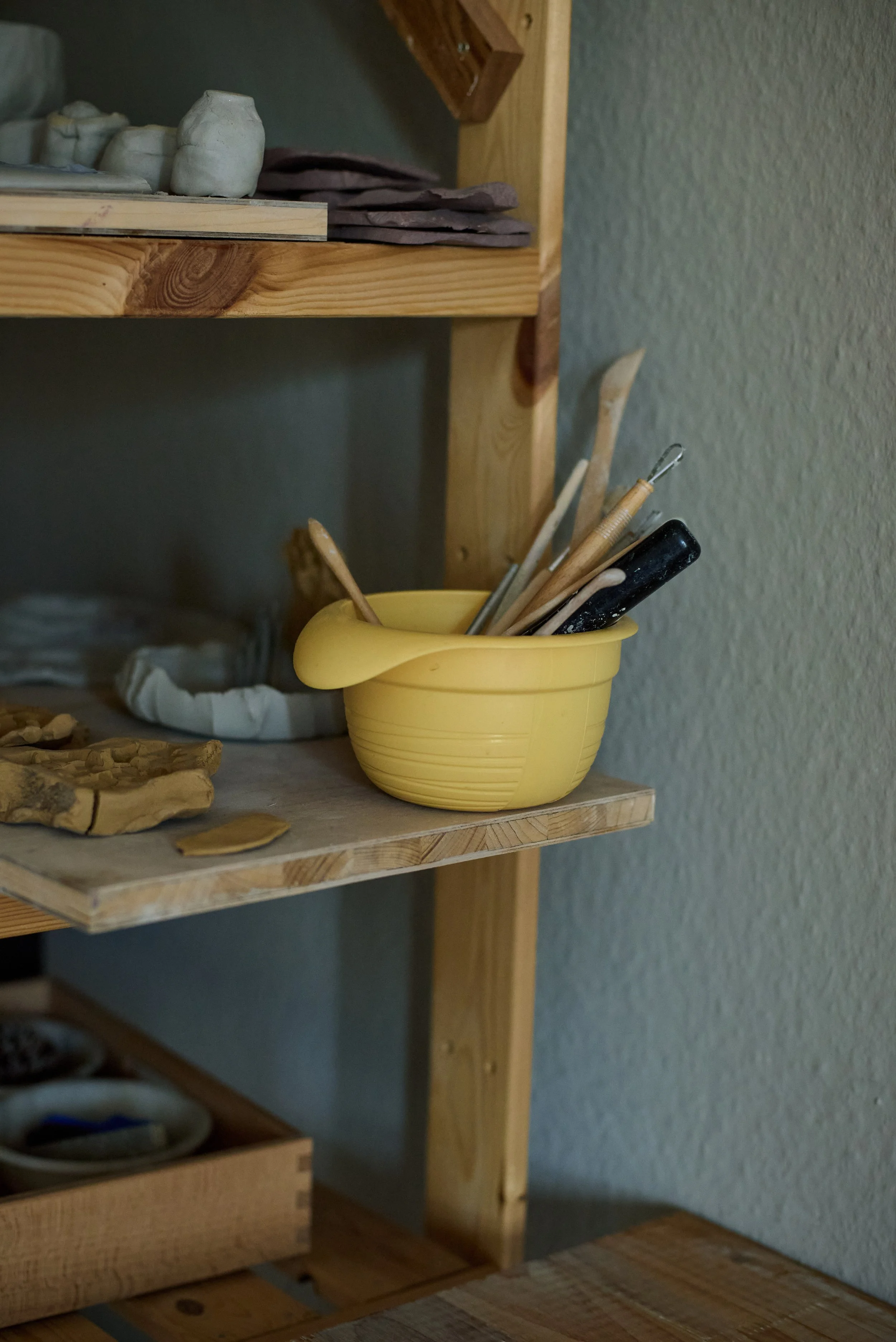 A yellow pottery bowl on a wooden shelf holding various pottery tools inside.