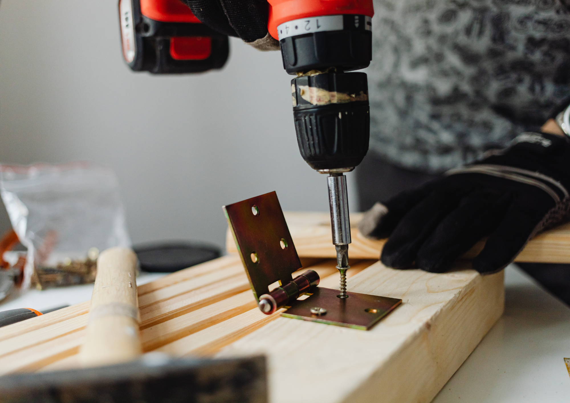 Une personne utilisant une perceuse électrique pour fixer une pièce métallique sur une planche en bois. La personne porte des gants noirs.