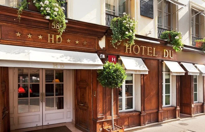 Façade en bois d'un hôtel avec des fleurs vertes en pot et en jardinière, enseigne en or 'Hôtel du', symbole 'HD' et un panneau rouge avec un H blanc.