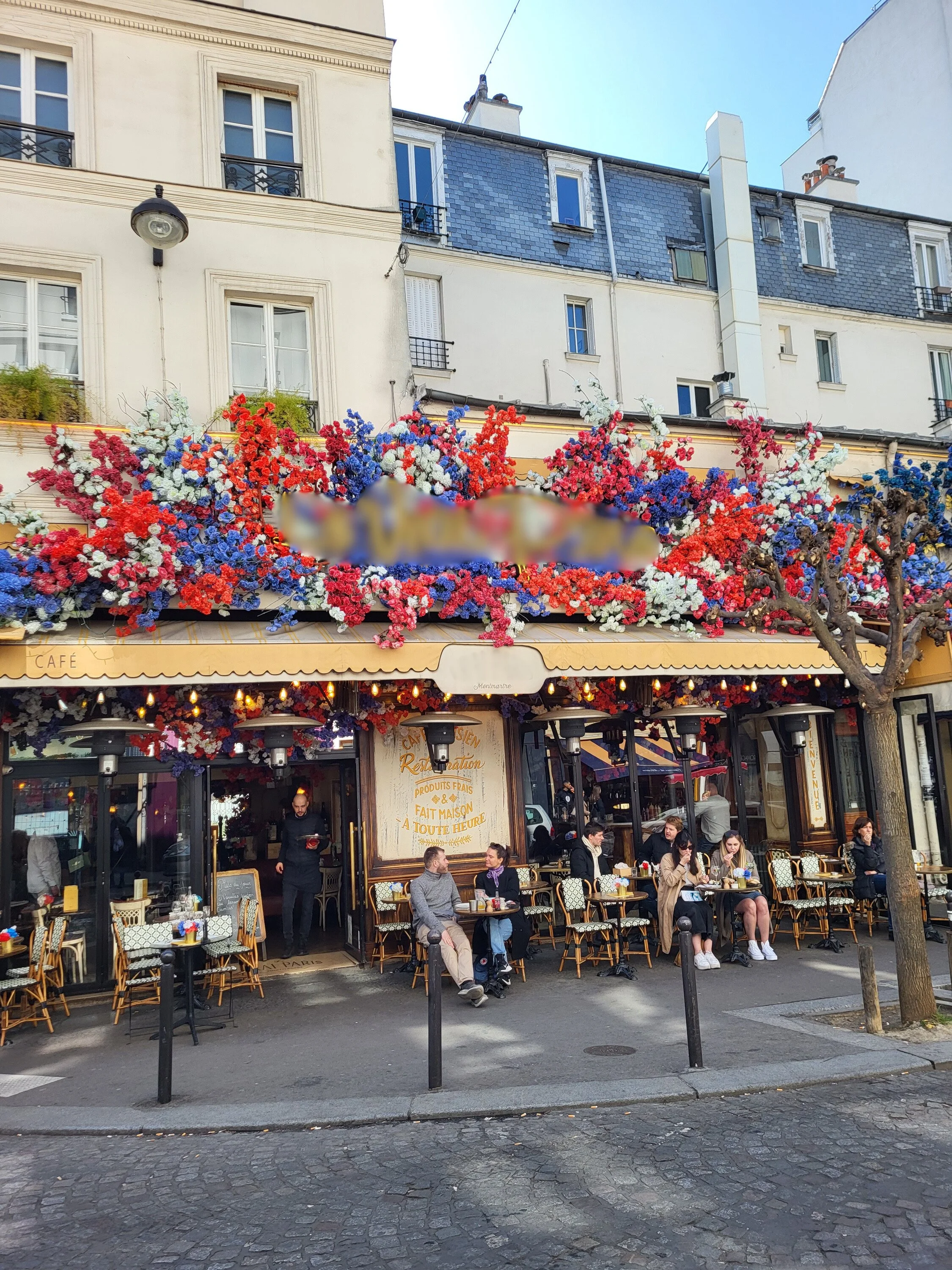 Café parisien avec terrasse extérieure, décorations florales colorées, plusieurs personnes assises à des tables, façade de bâtiment typique, arbres et pavés dans la rue.