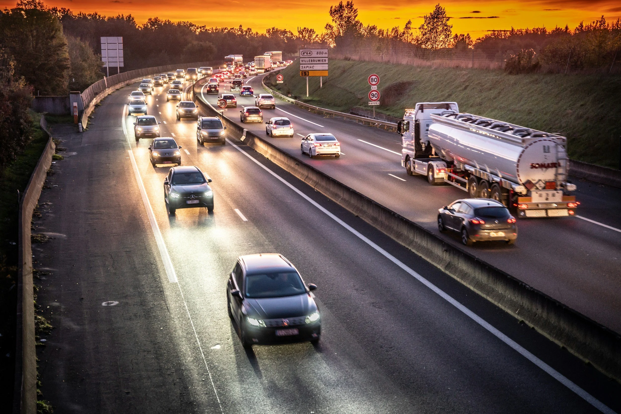 Une route à plusieurs voies en soirée ou au coucher du soleil. Beaucoup de voitures en circulation, avec un camion transportant un réservoir. La route est bordée d'une barrière de sécurité. Signes de limitation de vitesse et panneaux de direction sont visibles.