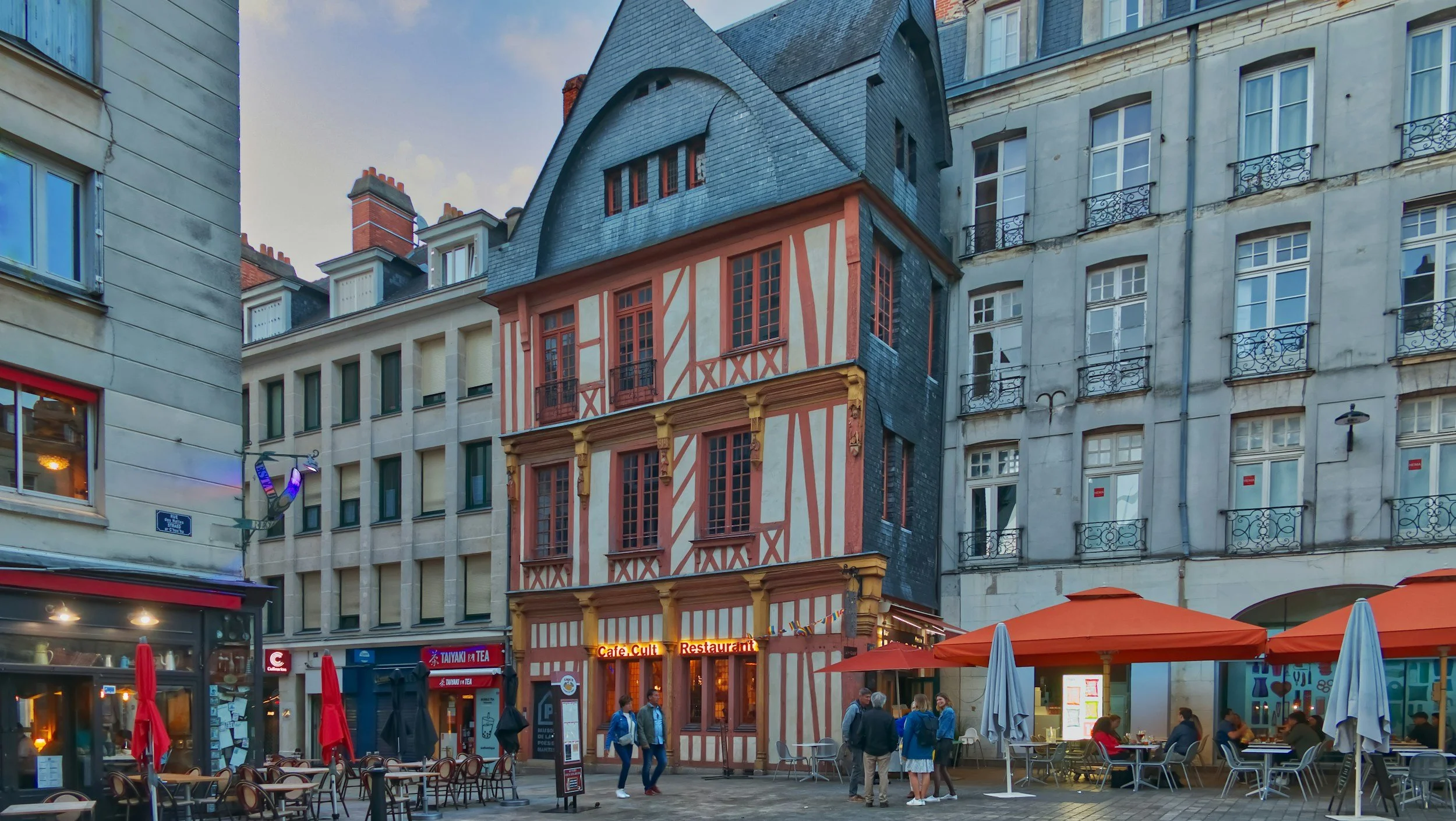 Une rue pavée avec une vieille maison à façades en bois rouge et beige, entourée de bâtiments modernes, avec des personnes assises et des tables à l'extérieur sous des parasols orange.