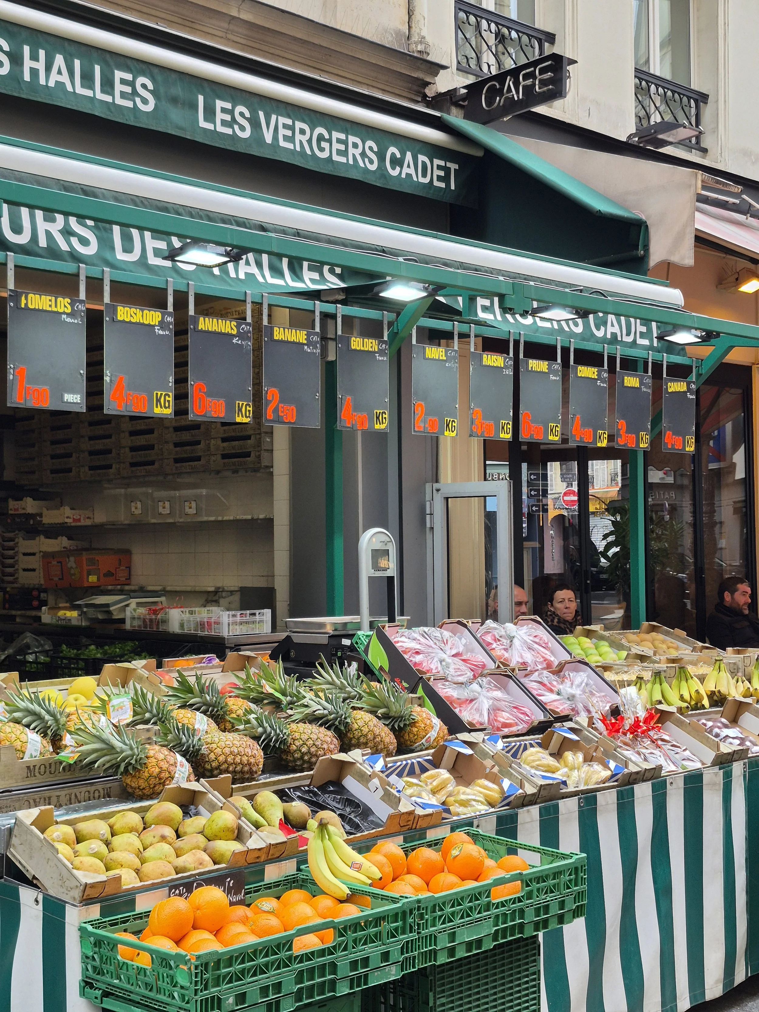 Stand de fruits frais avec ananas, oranges, bananes, pommes, et autres fruits, à l'extérieur d'un marché ou d'un café.