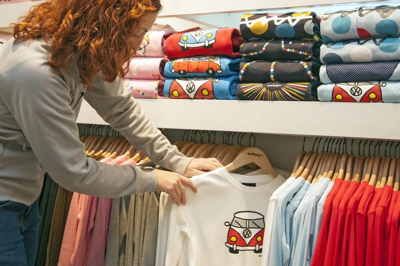 Une femme fait du shopping dans une boutique de vêtements, avec des t-shirts affichant des motifs de Volkswagen, notamment une van rouge et d'autres voitures colorées.