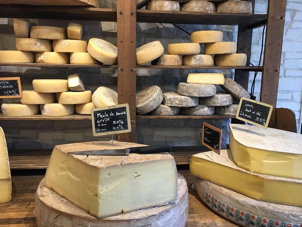 Différents types de fromages en train de vieillir, certains en rond, d'autres en blocs, exposés sur des étagères en bois, avec des étiquettes indiquant leurs noms et prix.