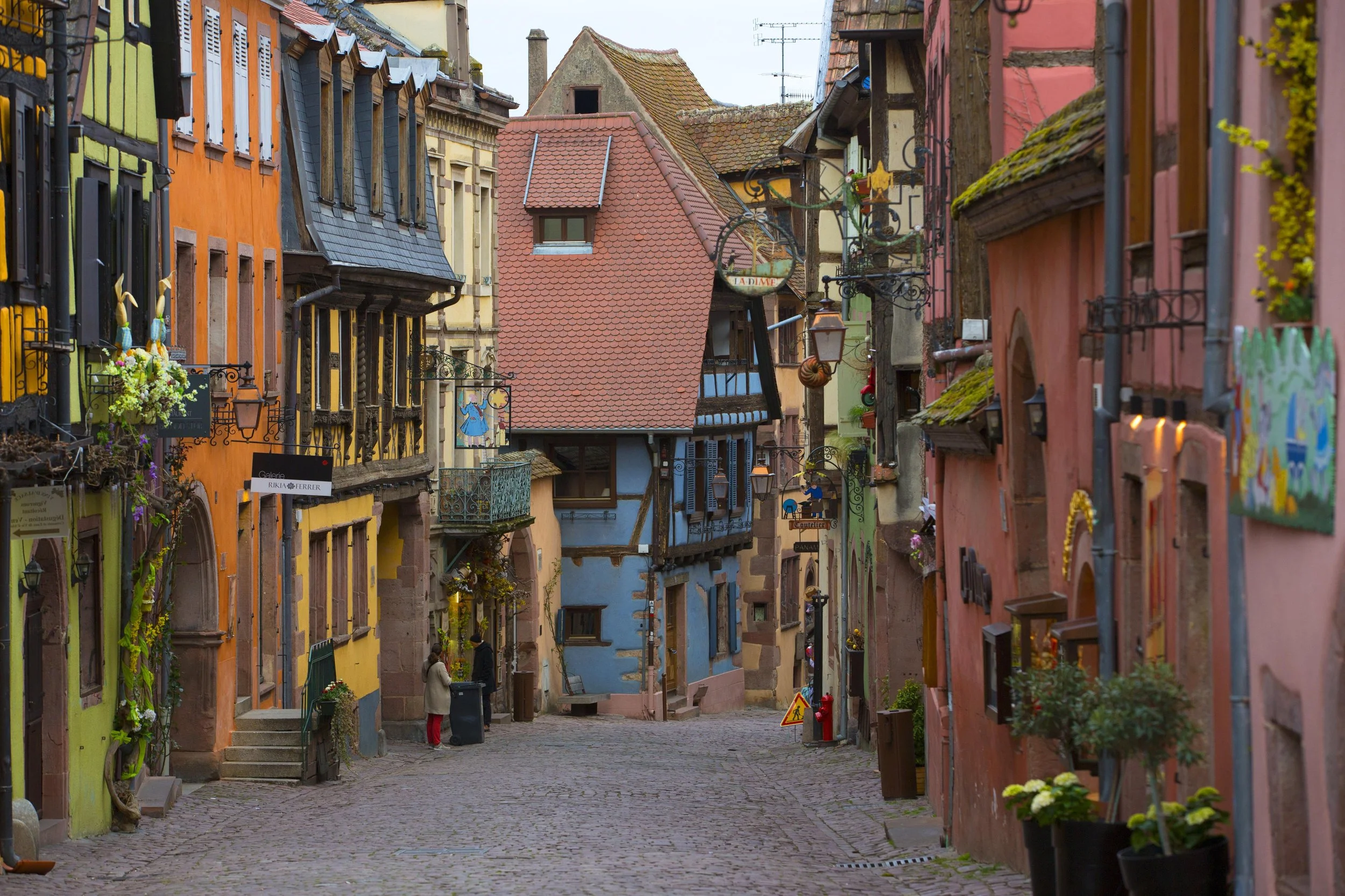 Rue pavée dans un village avec maisons colorées aux façades peintes, fenêtres ornées de fleurs, lampadaires et décorations suspendues.