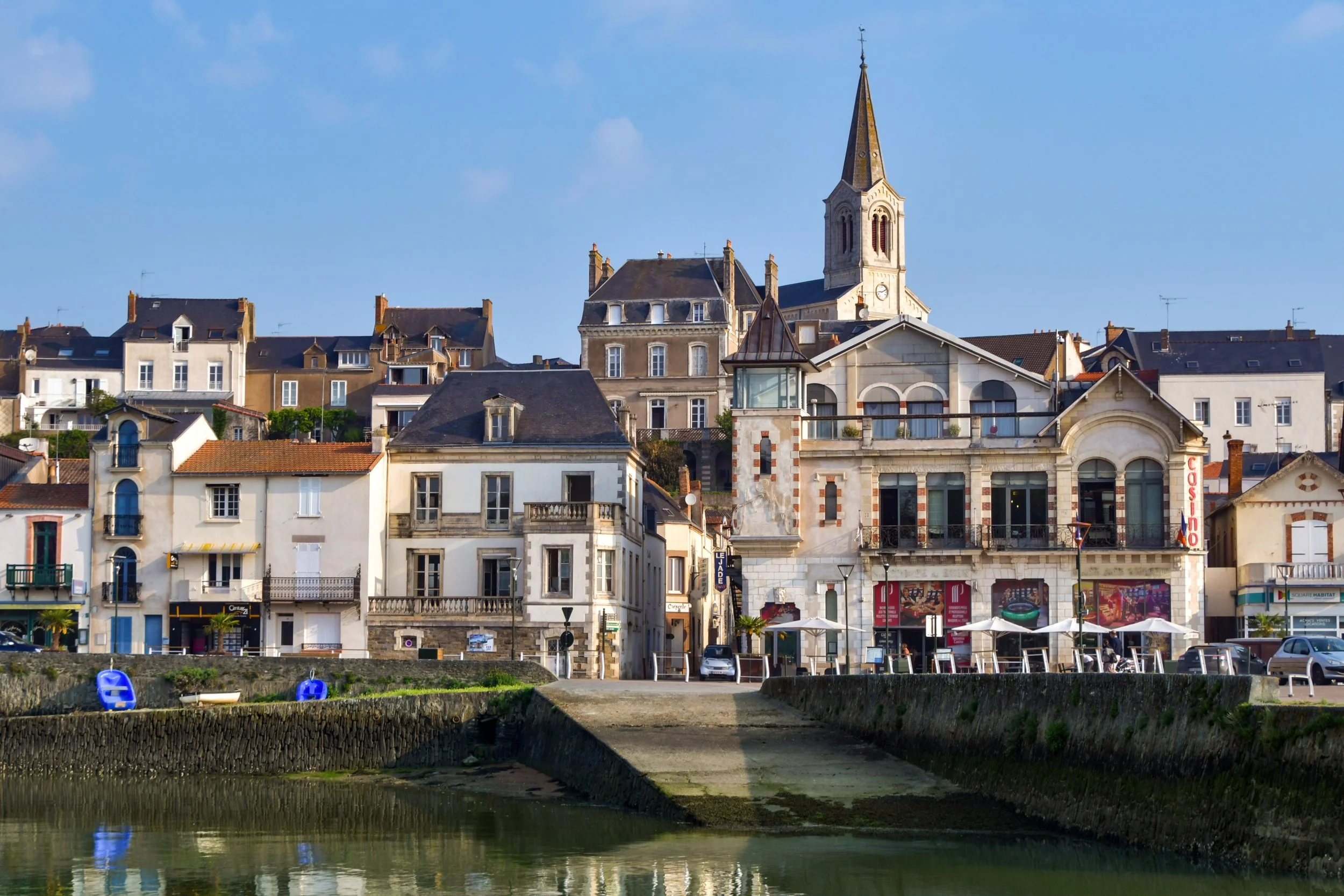 Vue d'un village côtier avec des bâtiments historiques, une église avec un clocher pointu, et des terrasses avec parasols en bord de rivière.