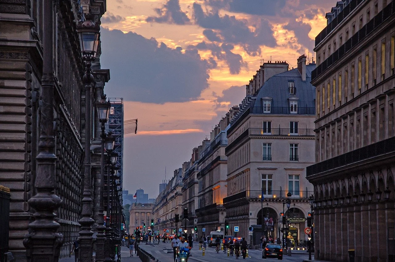 Vue d'une rue urbaine parisienne au crépuscule avec des bâtiments haussmanniens, des lampadaires et des véhicules en circulation.