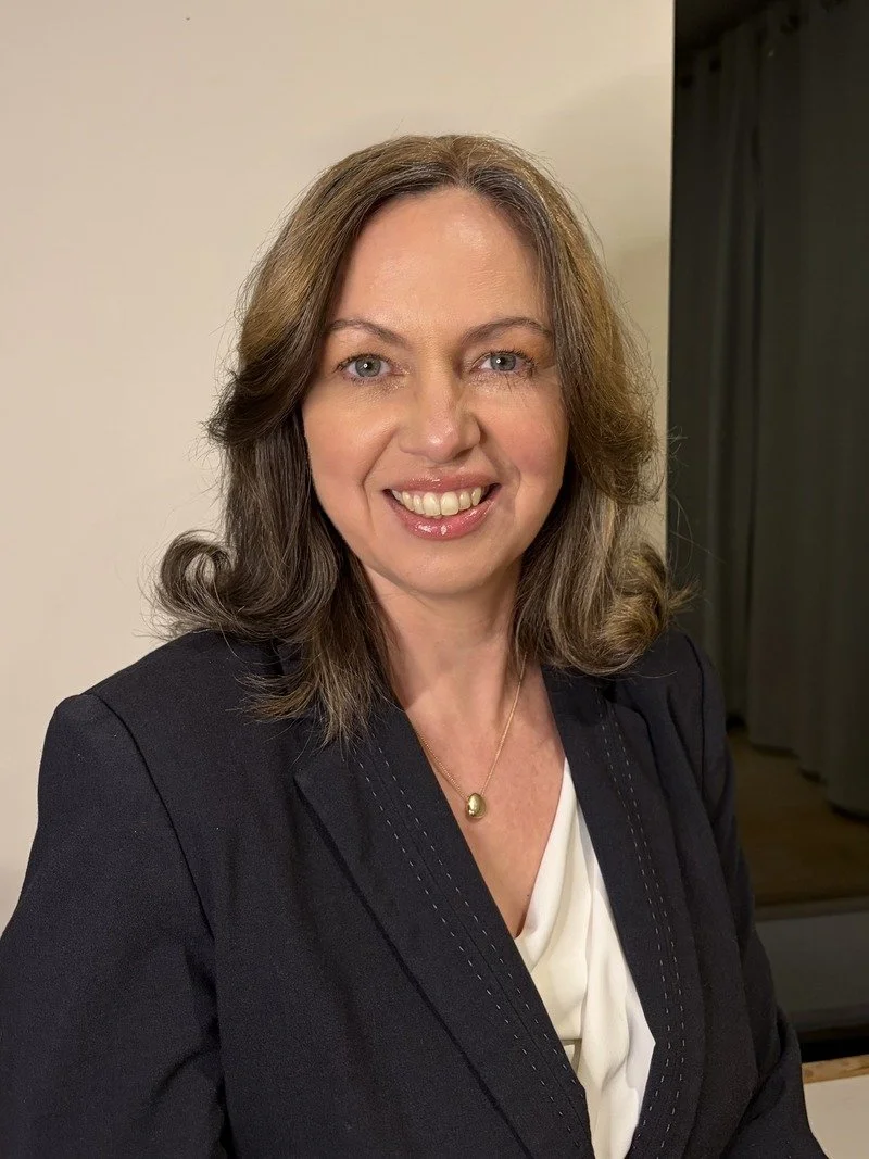 Amanda Mugnier, Edvance Recruitment founder. Shoulder-length wavy brown hair, wearing a black blazer with a white top underneath and a gold necklace with a pendant, smiling at the camera in an indoor setting.