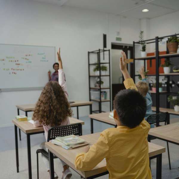 photo taken from behind with students sat and their desks with hands in the air answering their teacher
