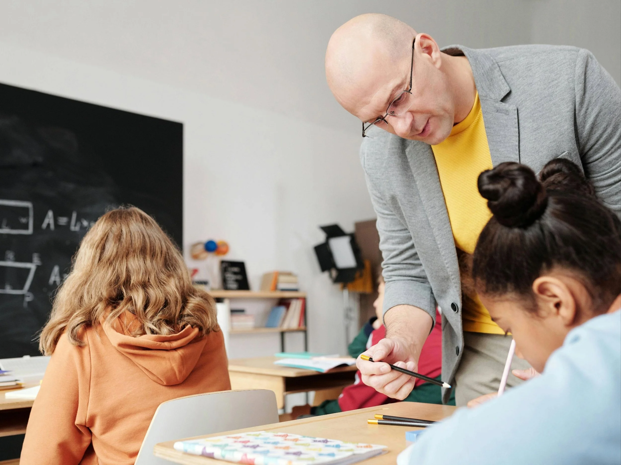 A male teacher with glasses and a grey blazer helping students in a classroom with a blackboard and books, engaging with a girl with dark hair in a bun.