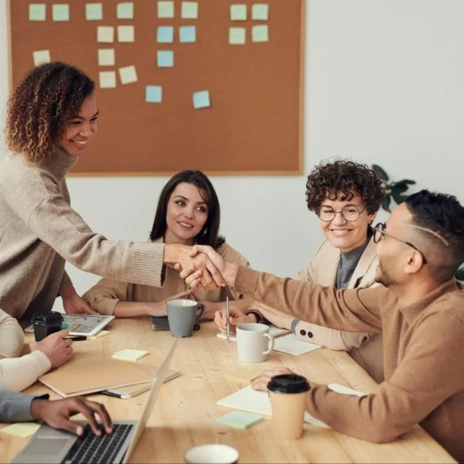 Woman stood up shaking man's hand around a meeting table with other colleagues smiling