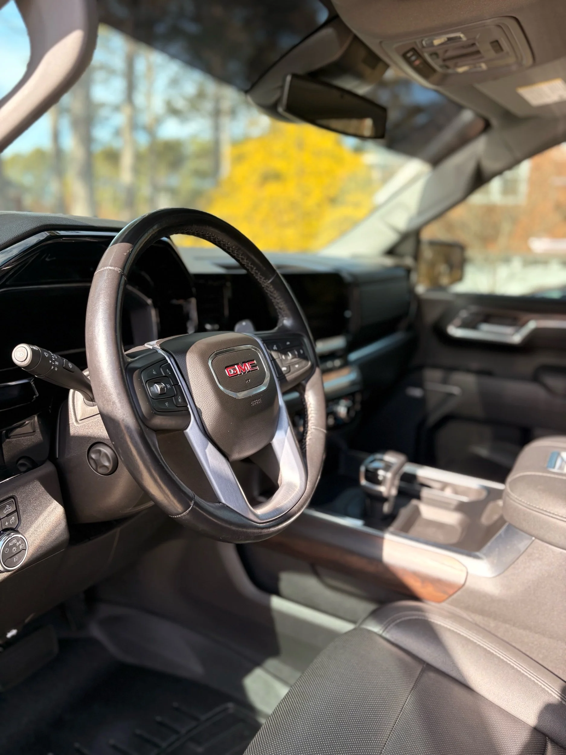 Interior of a GMC vehicle, showing the steering wheel, dashboard, gear shift, and part of the passenger seat, with trees visible through the windshield.