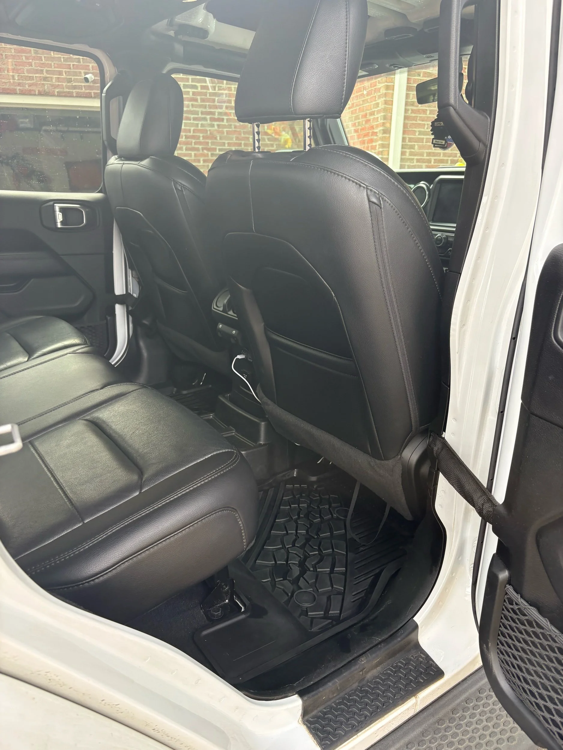 Interior of a vehicle showing black leather seats, the back of the front seats, and the rear seats with black rubber floor mats. The vehicle has a white exterior, visible from the door opening, and a brick wall outside.