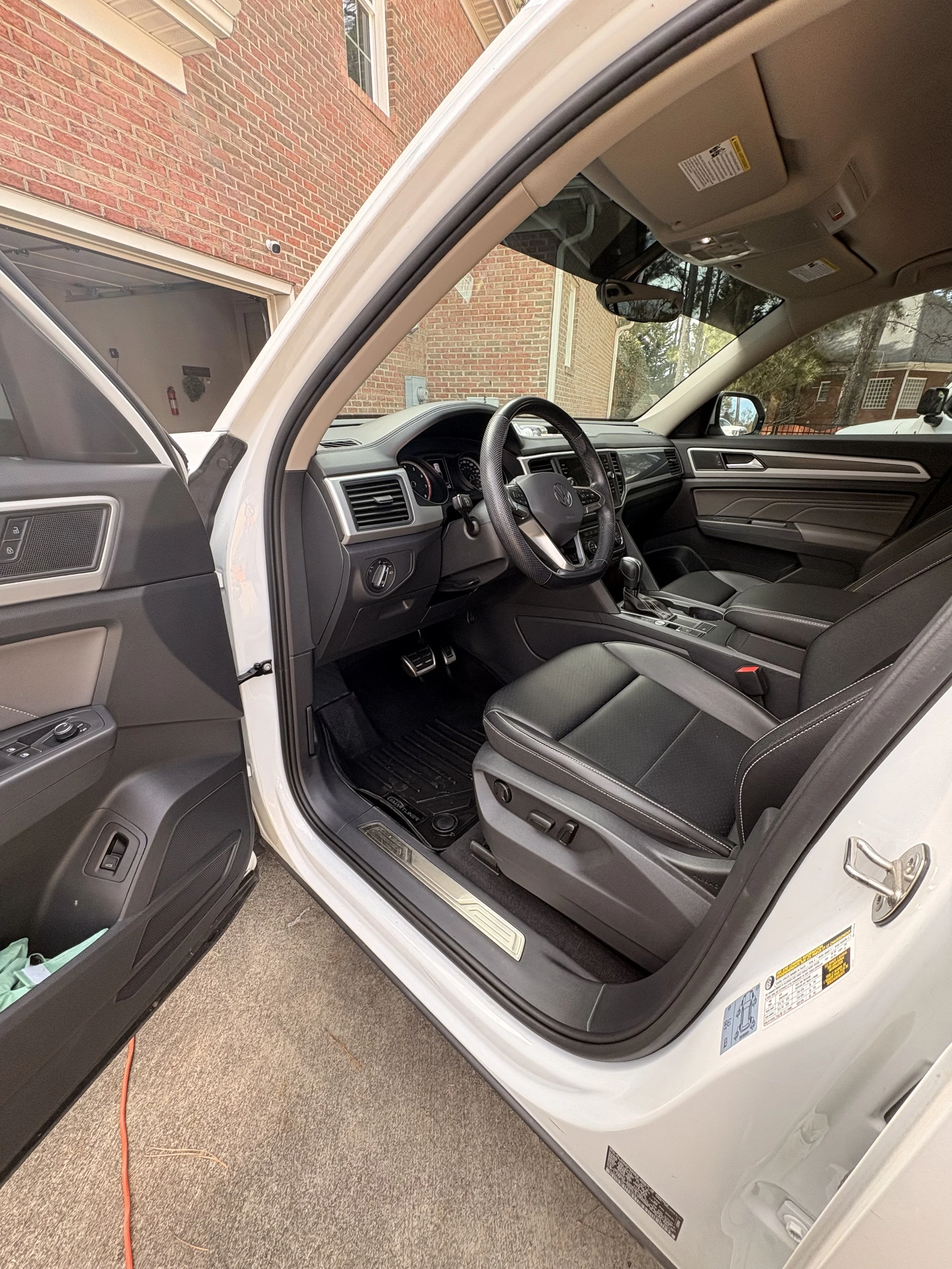 Interior of a white vehicle's front seat area, showing the driver's seat, dashboard, steering wheel, and part of the front passenger seat, with a brick house exterior visible through the windshield and door.