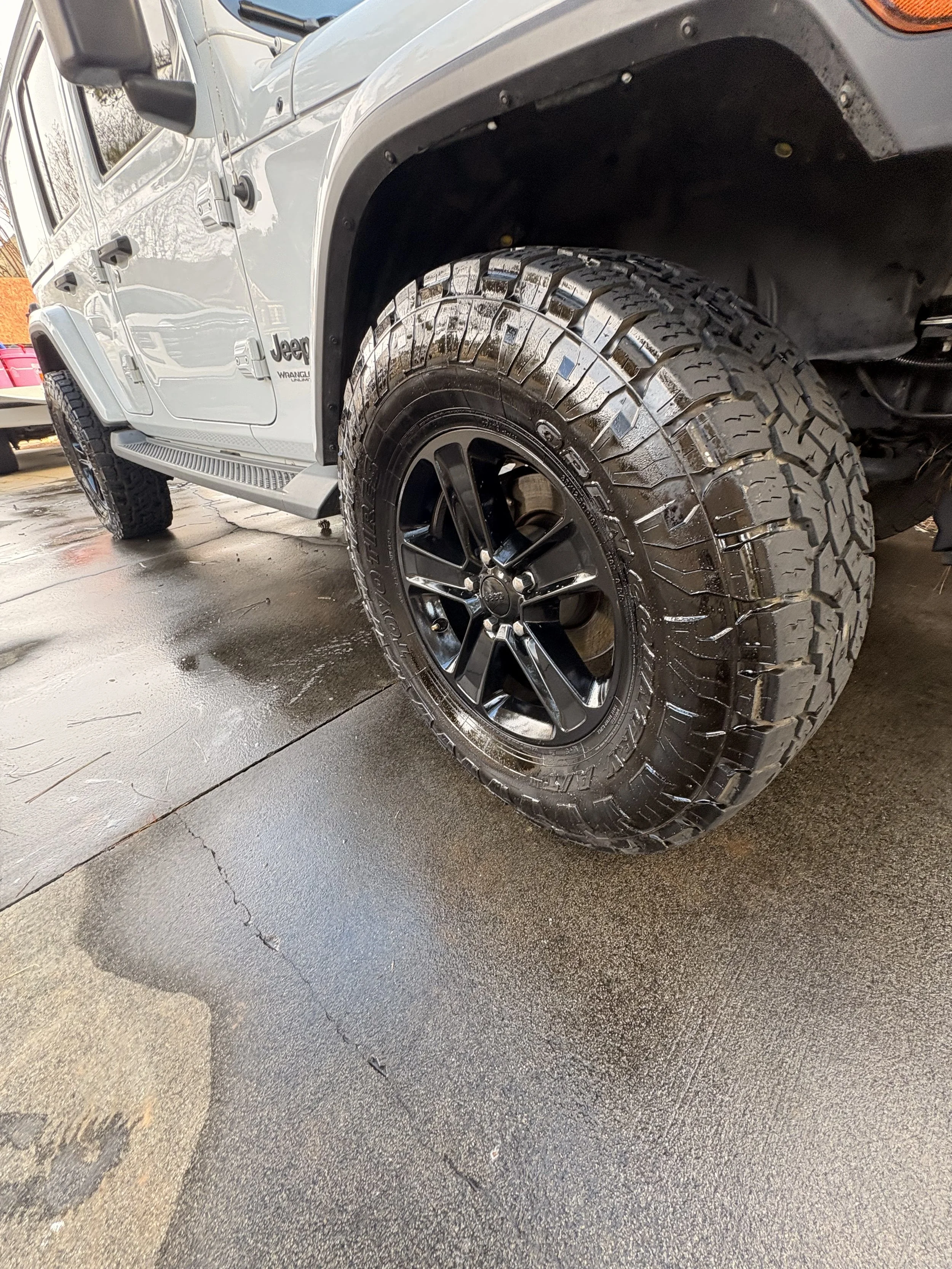 Close-up of a Jeep vehicle's front tire and wheel, with a black rim and rugged off-road tire, parked on a wet concrete surface.