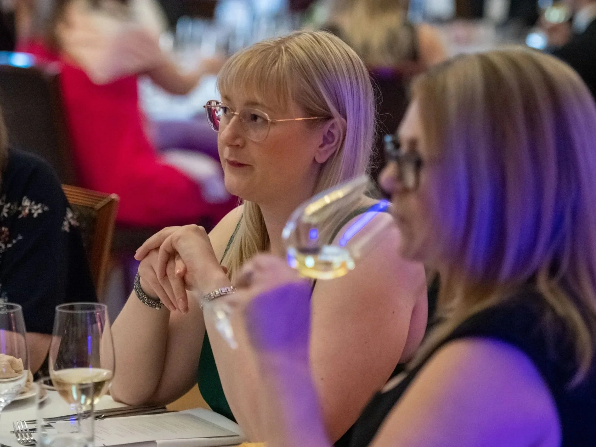 A woman with blonde hair and glasses is sitting at a dinner table during a social event, looking to the side with her hands clasped in front of her. There are wine glasses and plates in front of her, and other people are visible in the background.