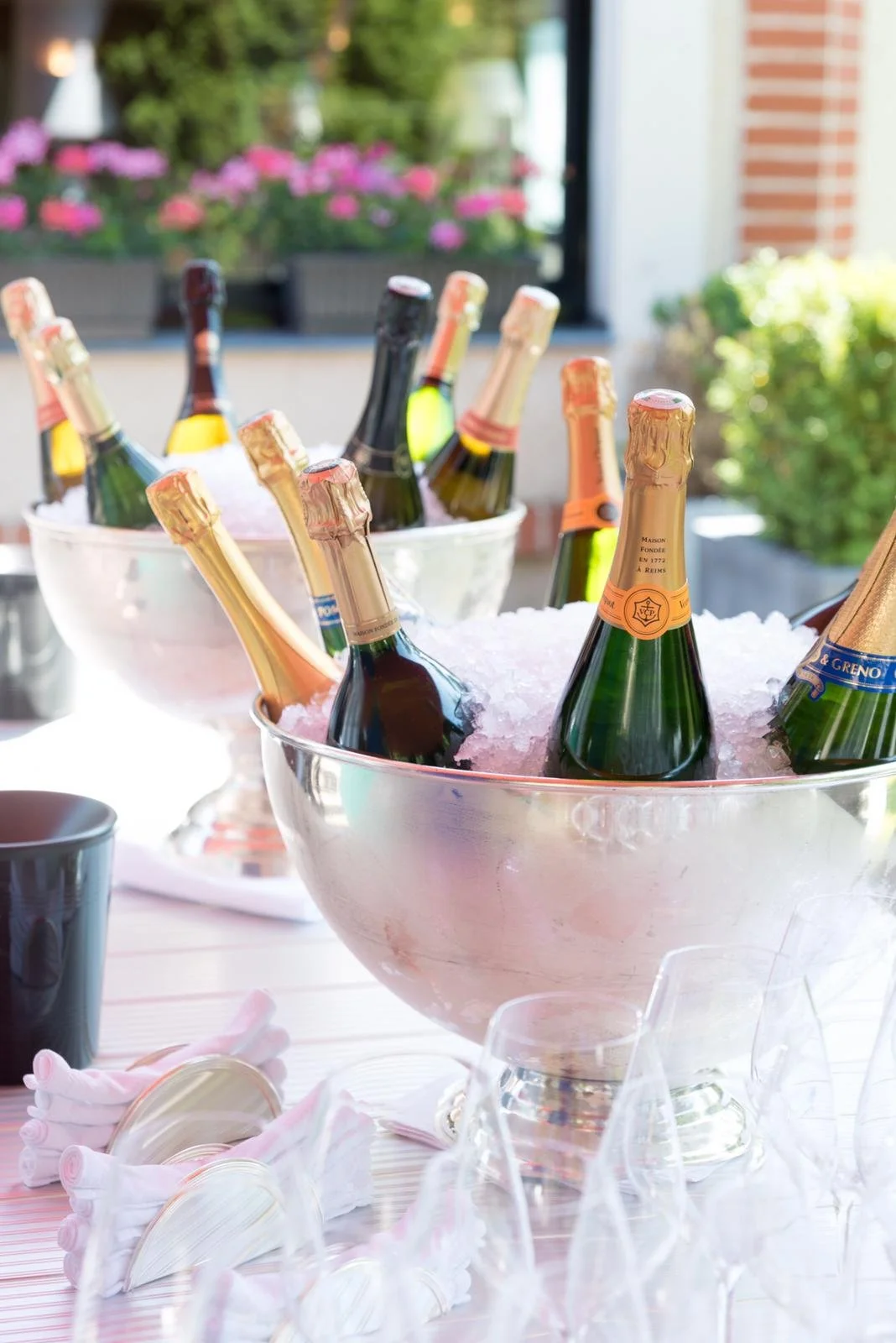 Champagne bottles chilling in ice buckets on a table, with glassware and pink napkins nearby, outdoors setting with flowers and greenery in background.