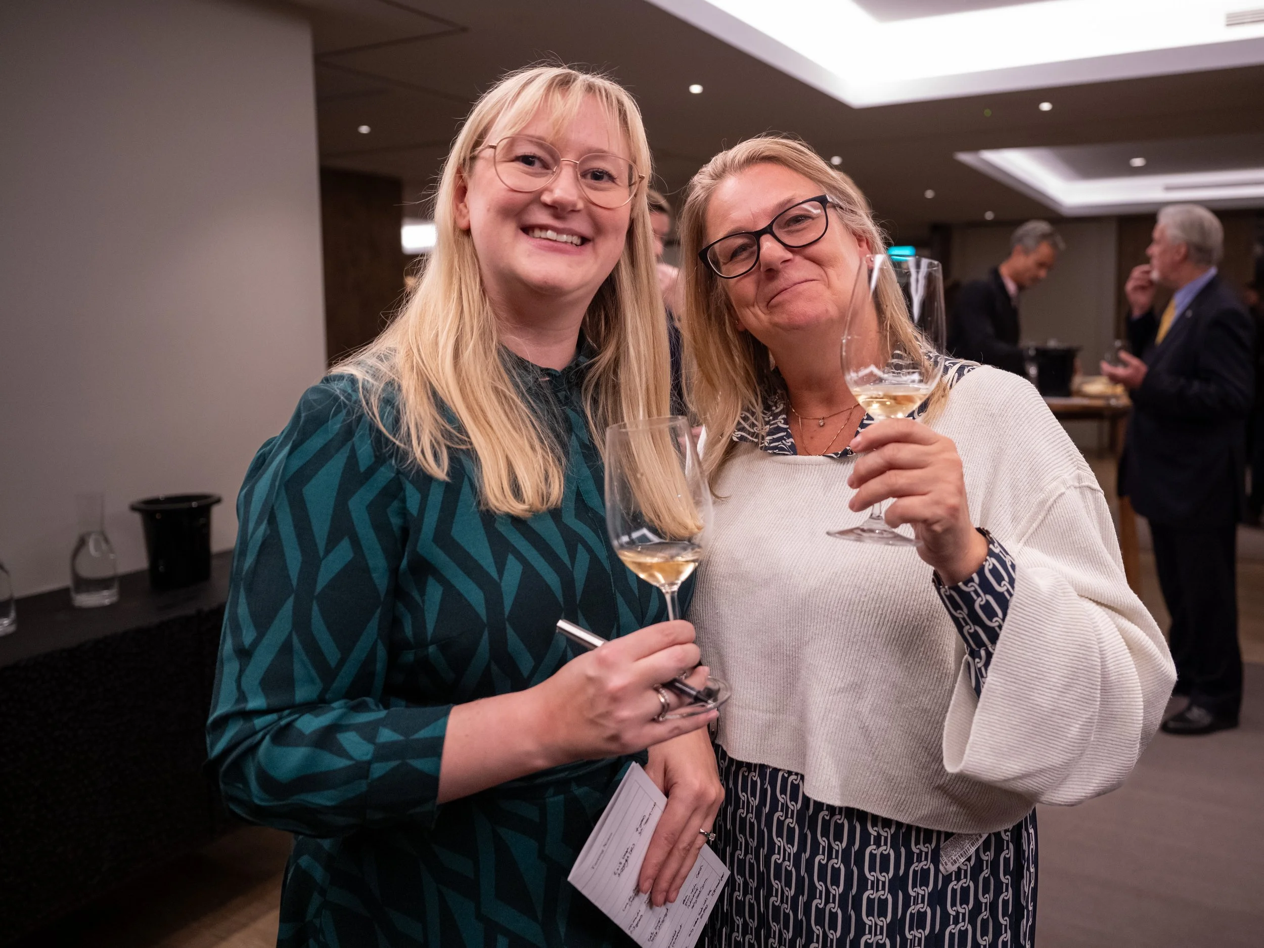 Two smiling women holding wine glasses at a social event, with other people in the background.