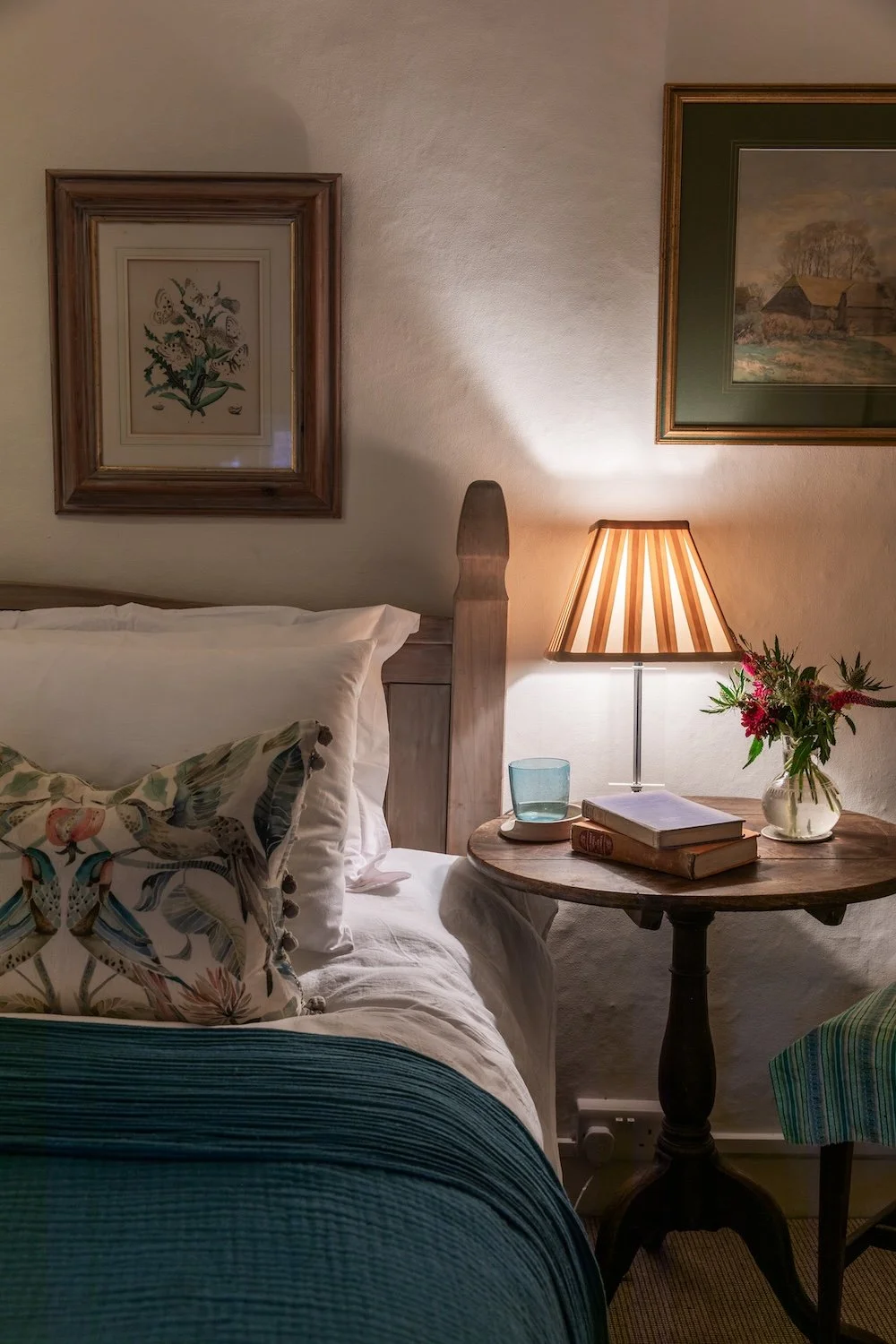 A cozy bedroom corner with a wooden bed, decorative pillows, a round wooden side table, a striped lampshade, a small vase with flowers, and framed artwork on the wall.