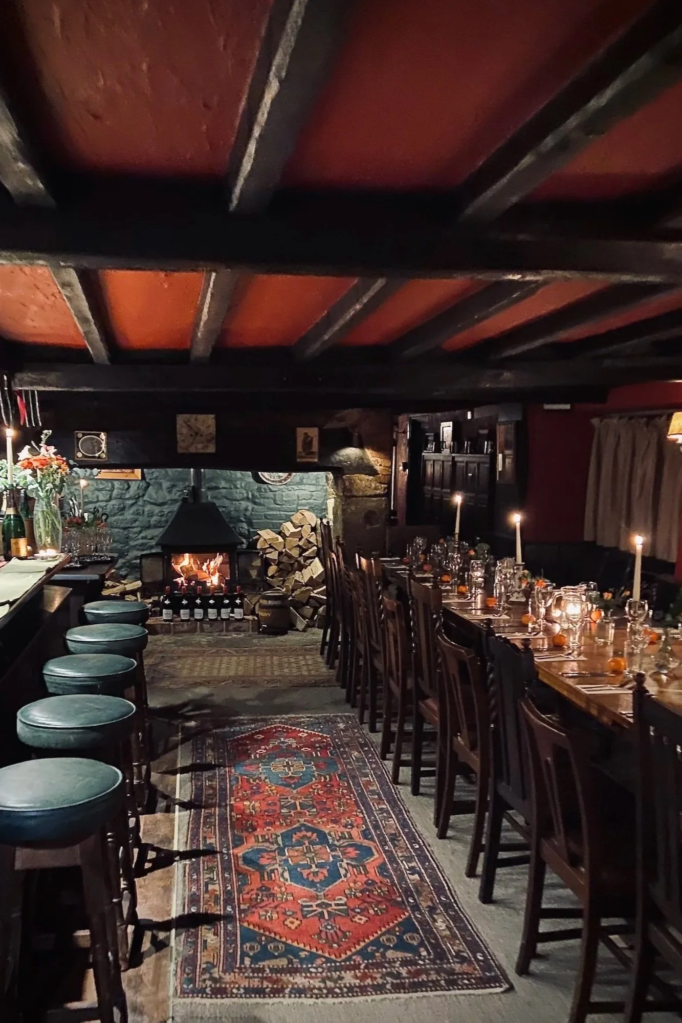 A cozy, dimly lit dining room with a long wooden table set with glasses, candles, and small pumpkins, and a fireplace with stacked logs in the background.