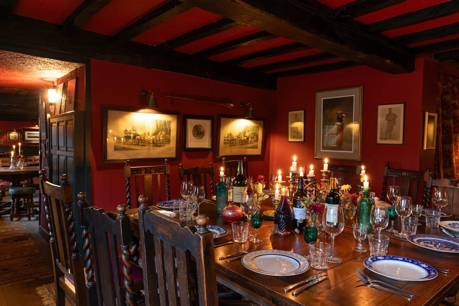 A dining room with a long wooden table set for a meal, decorated with candles, bottles of wine, glasses, and plates, with framed artwork on red walls and dim lighting.