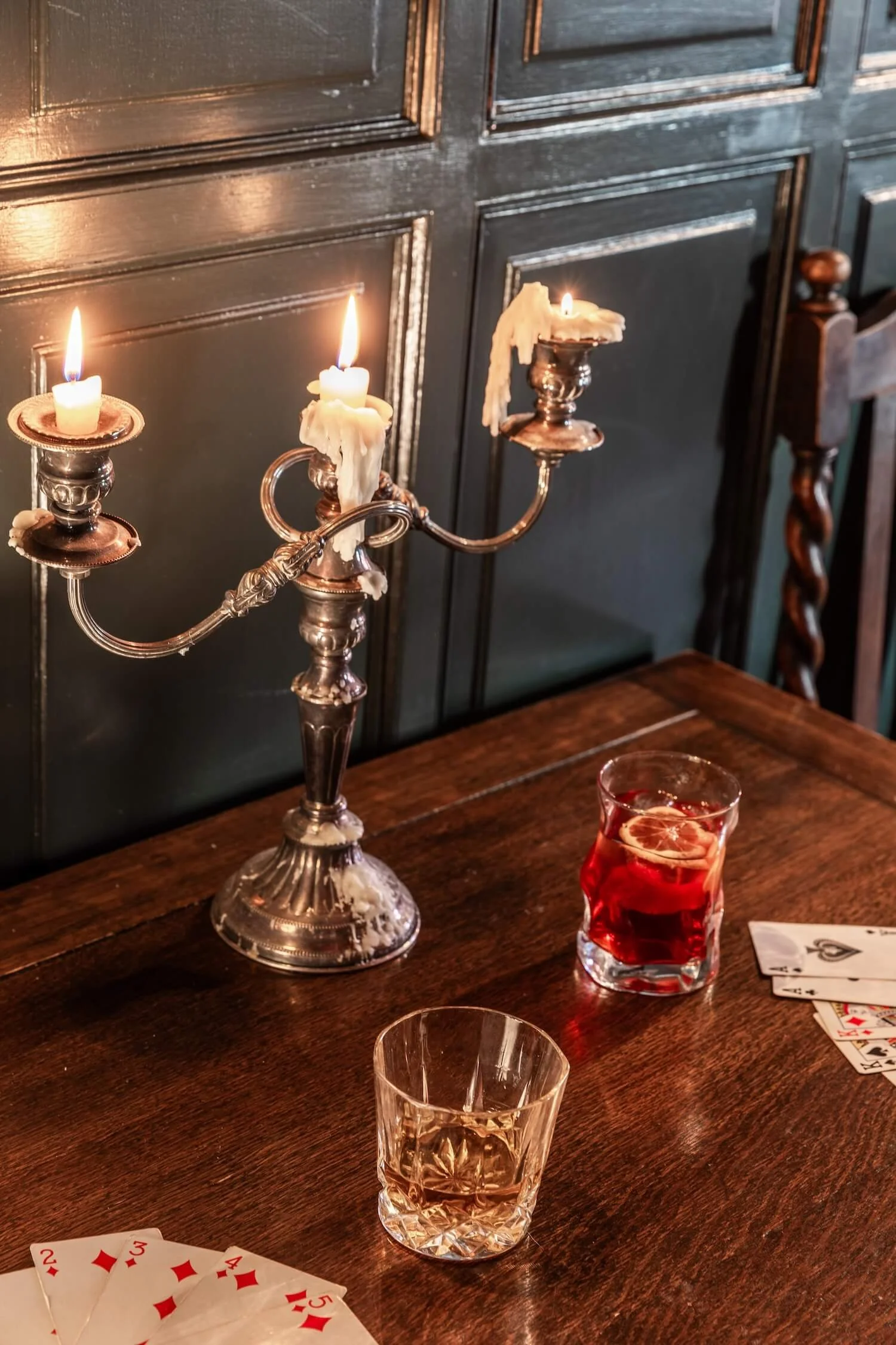 A wooden table with a vintage candelabra holding three lit candles, two glasses of drink (one with lemon and one with a slice of lime), and playing cards spread out on the right side, against a dark paneled wall background.