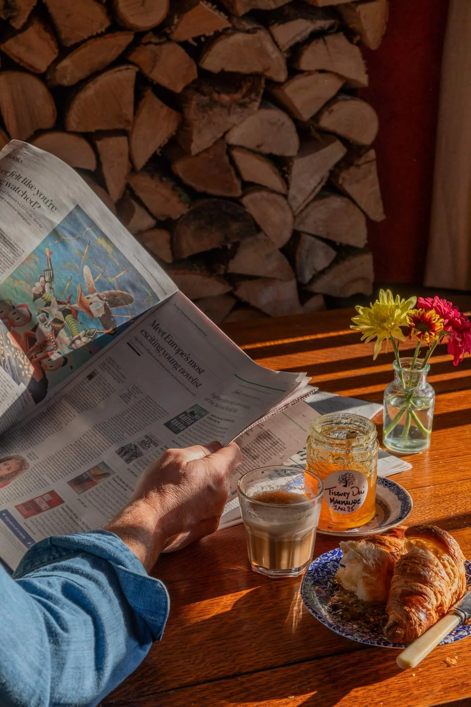 A person reading a newspaper at a wooden table with a croissant, a glass of coffee, a jar of marmalade labeled 'Tisbury Dev Marmalade', and a small vase with yellow and pink flowers, with a woodpile in the background.