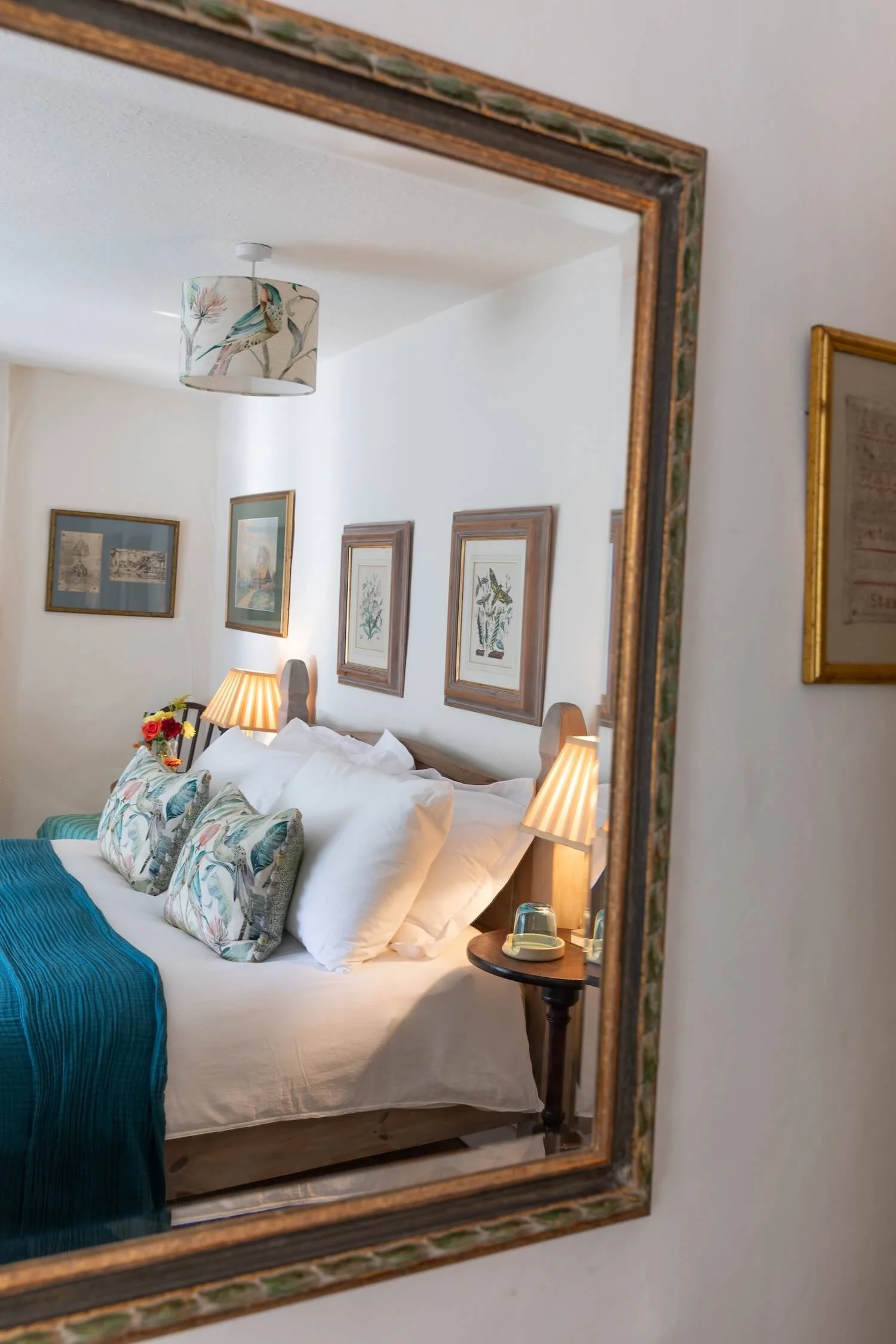 Reflection in a mirror shows a cozy bedroom with a bed, decorative pillows, a teal bedspread, wooden headboard, wall art, lamps, and small bedside tables.