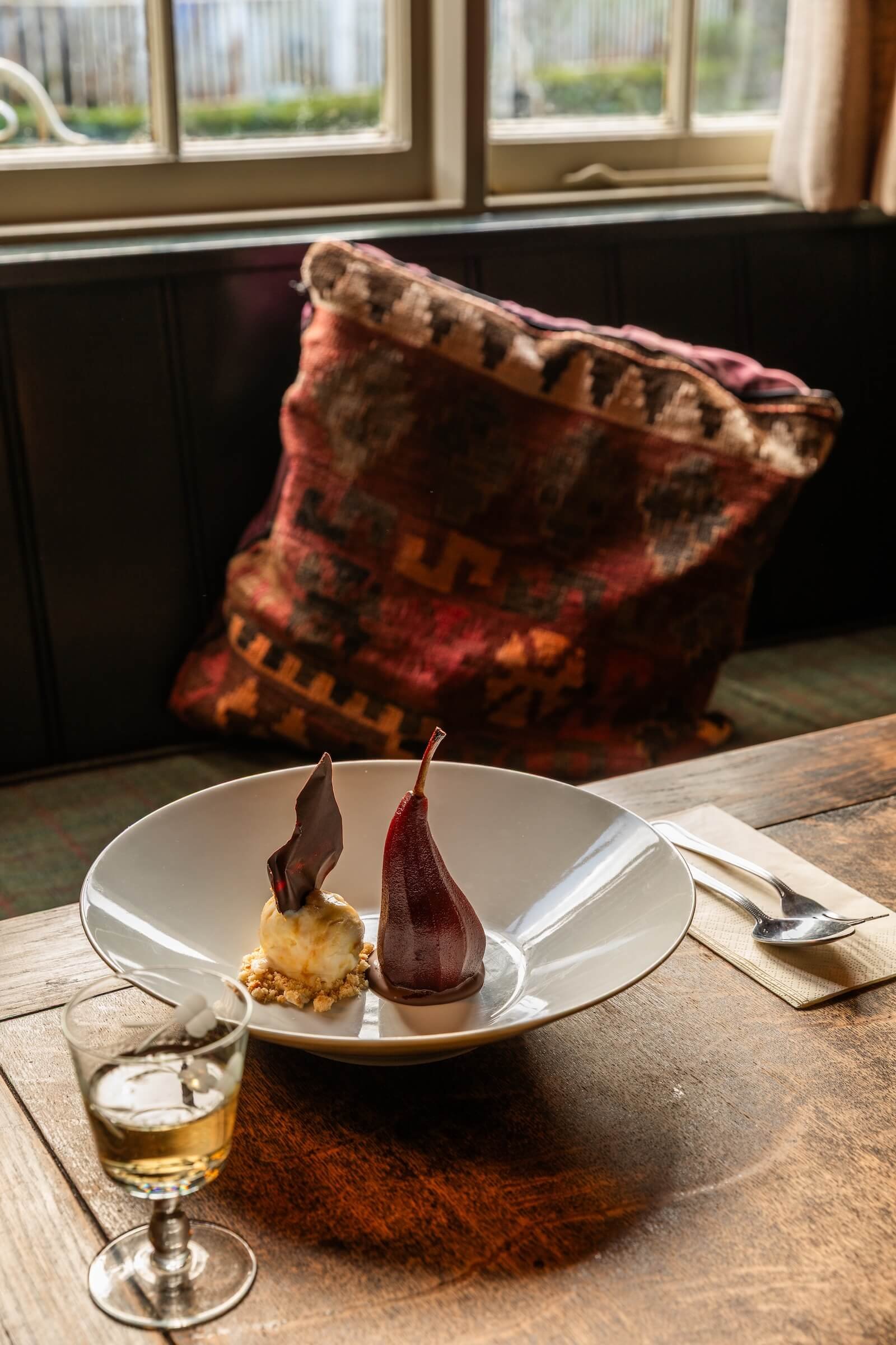 A dessert plate with two artistic chocolate garnishes, a scoop of ice cream with a chocolate piece, and a glass of white wine on a wooden table, with a patterned cushion and window in the background.