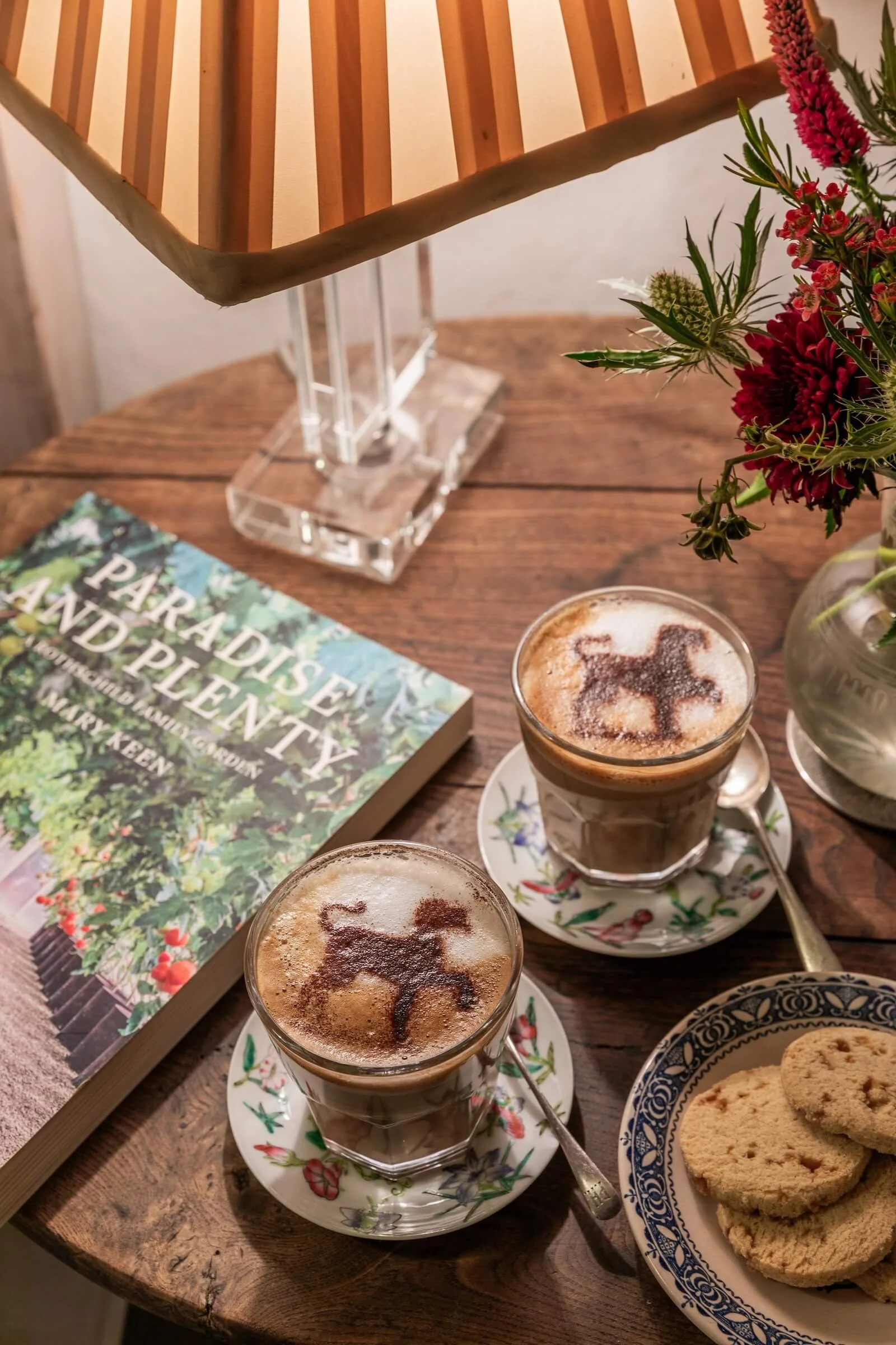 Two glasses of coffee with dog-shaped cocoa powder designs on top, on a wooden table with a book titled 'Paradise and Plenty,' a flower arrangement, and a plate of cookies.