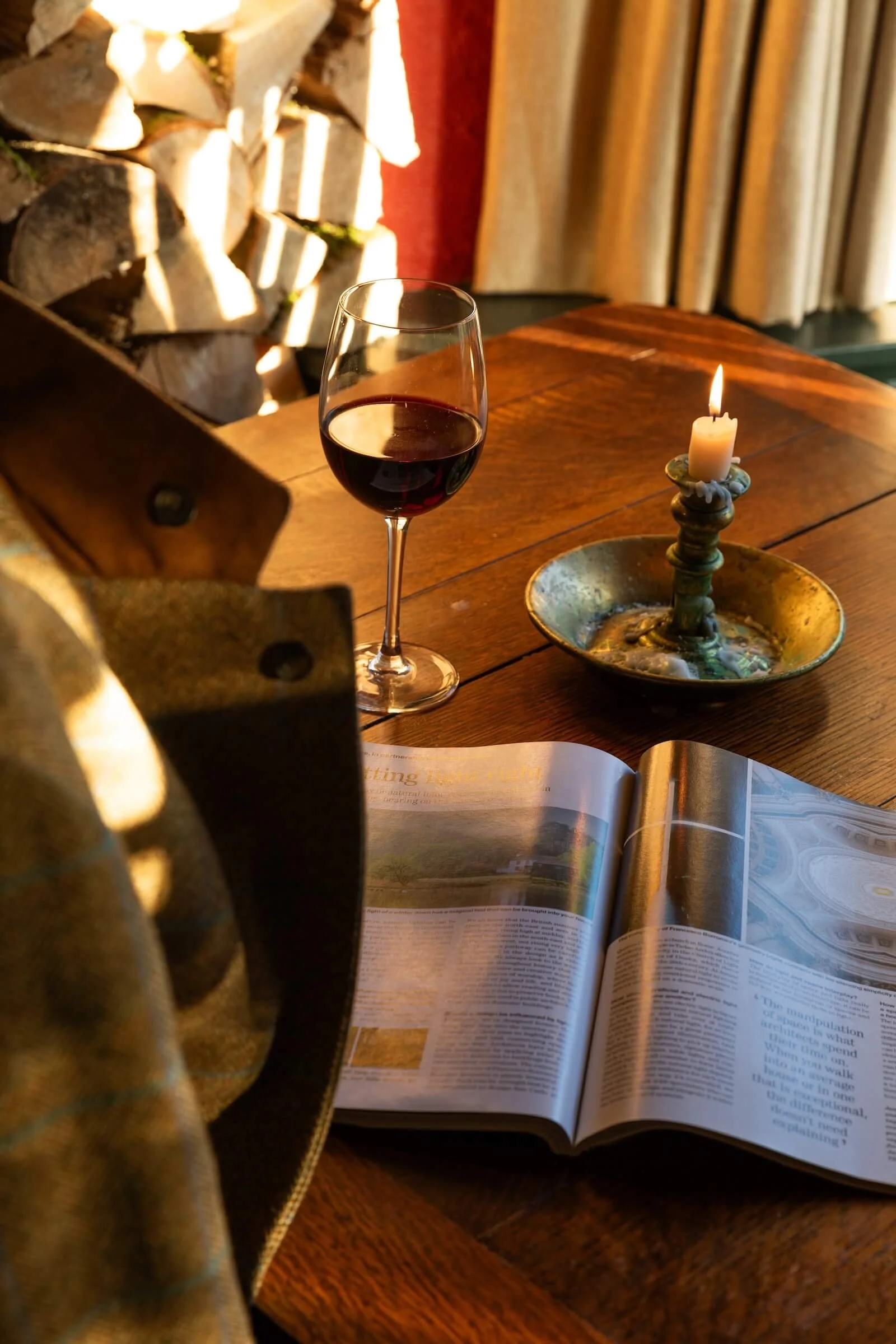 A wooden table with a glass of red wine, an open magazine, and a lit candle in a decorative holder, with a cozy indoor setting and a stack of firewood in the background.