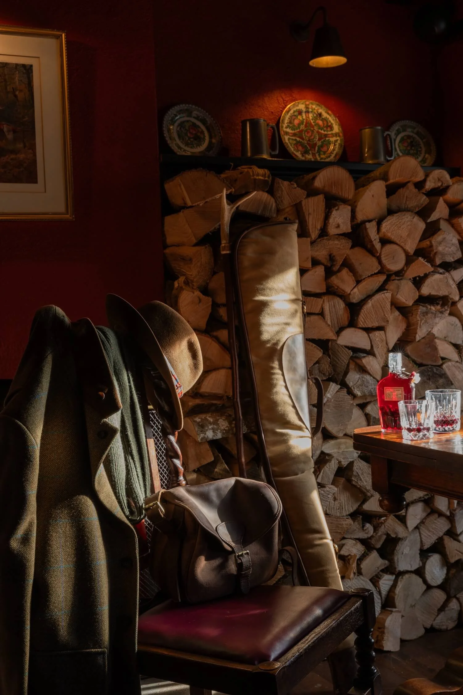 A cozy interior scene with a stack of firewood against the wall, a chair with a coat and a hat hanging on it, a bag resting on the chair, and a small wooden table with glasses and a red bottle, illuminated by warm lighting.