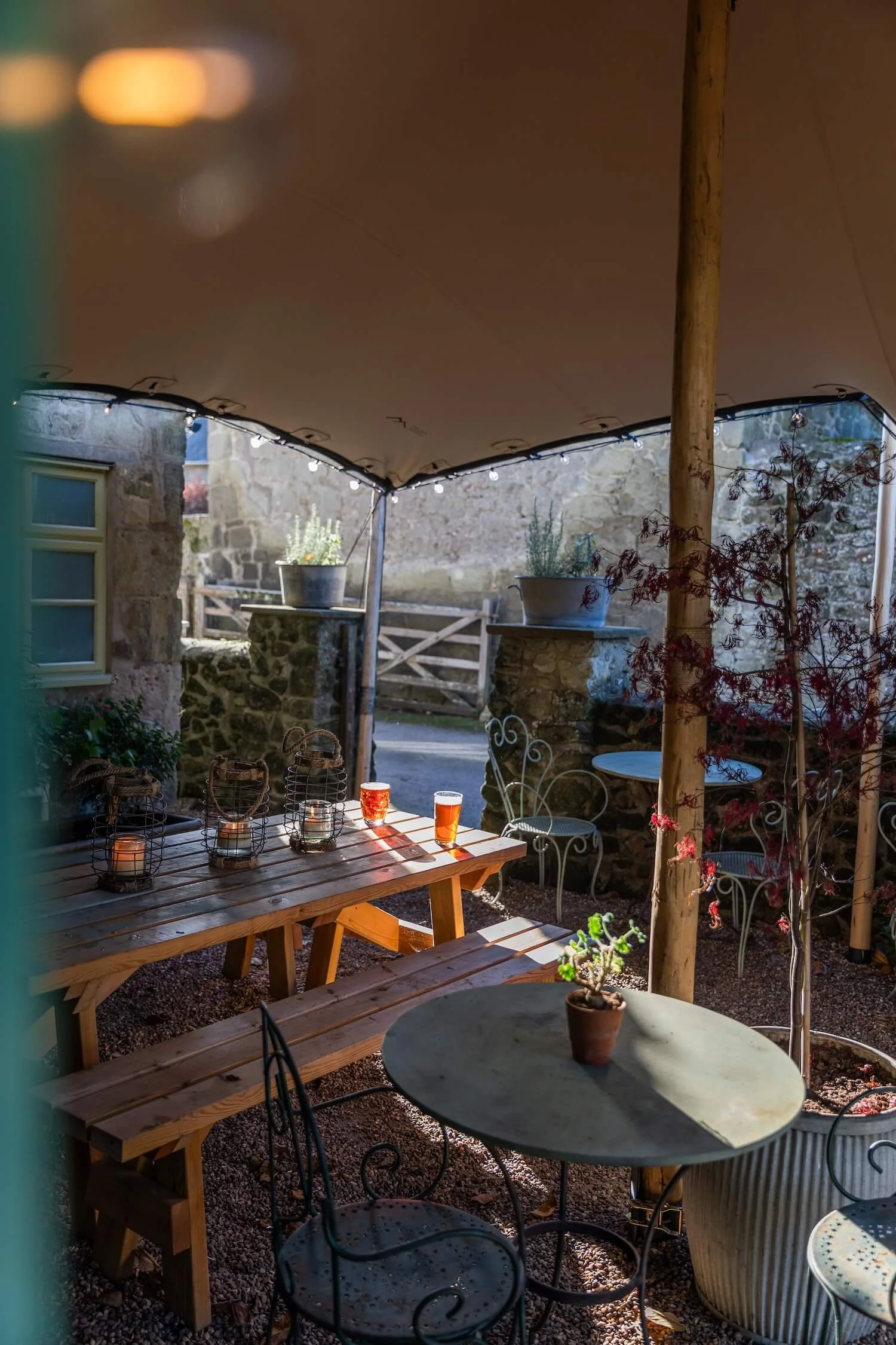 Cozy outdoor seating area with wooden and metal tables, potted plants, and string lights under a canopy or tent.