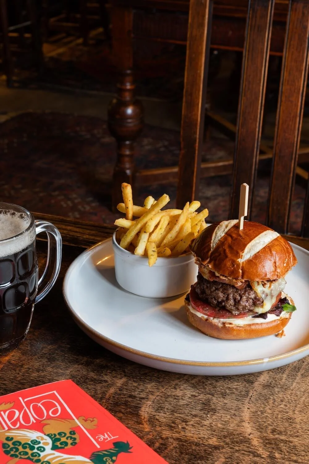 A plate with a cheeseburger, French fries, and a mug of dark beer on a wooden table in a restaurant setting.
