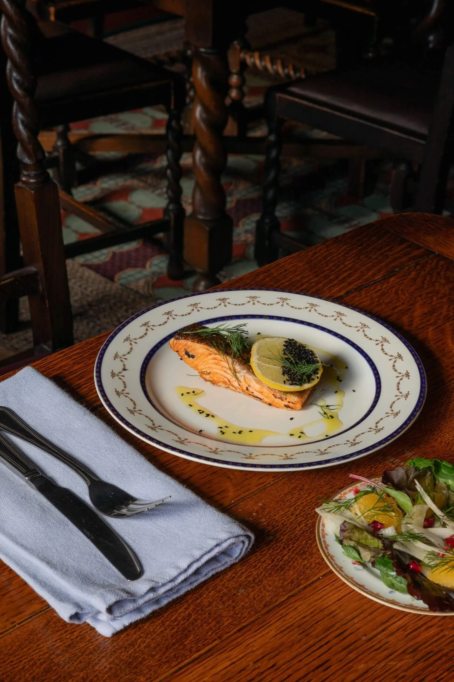 A plated piece of grilled salmon garnished with a lemon slice, herbs, and olive oil on a wooden table, alongside a small salad with greens and citrus slices.