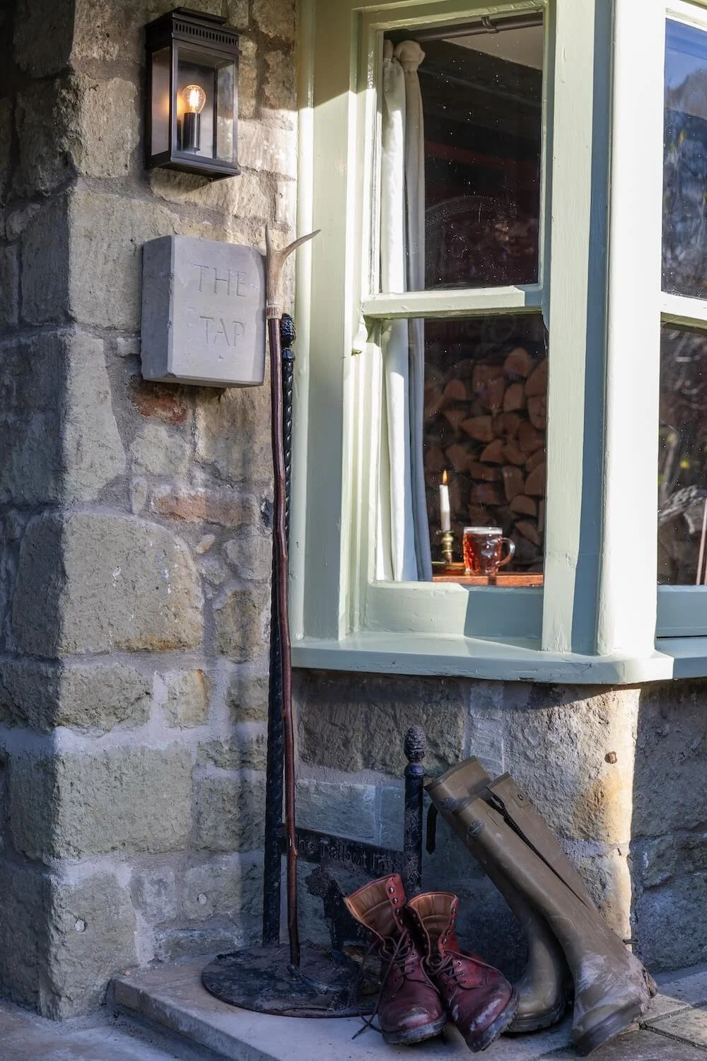 Close-up of a stone house wall with a black wall-mounted lantern, a white mailbox labeled 'THE TAP,' and a tall walking stick. In front, there's a pair of brown leather boots and a pair of tall rubber boots placed on the ground. The window shows a lit candle, a glass mug filled with a beverage, and a stack of firewood inside.