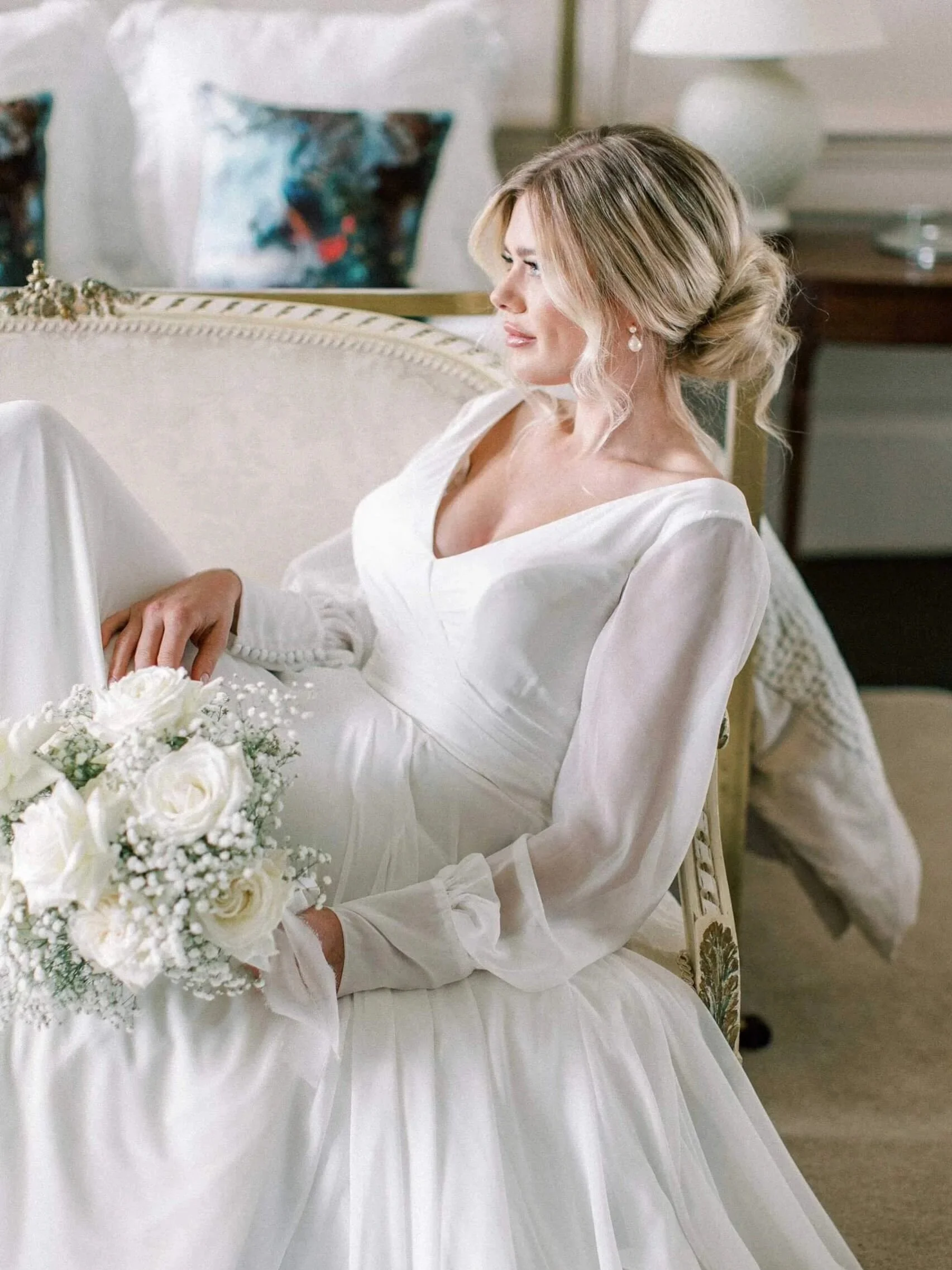 A woman dressed in white sitting on an ornate cream-colored couch with a bouquet of white flowers, with a decorative pillow and a side table with a lamp in the background.