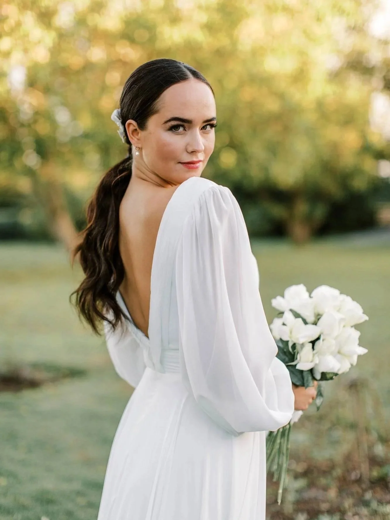 A woman in a white dress holding a bouquet of white flowers outdoors with a background of trees and greenery.