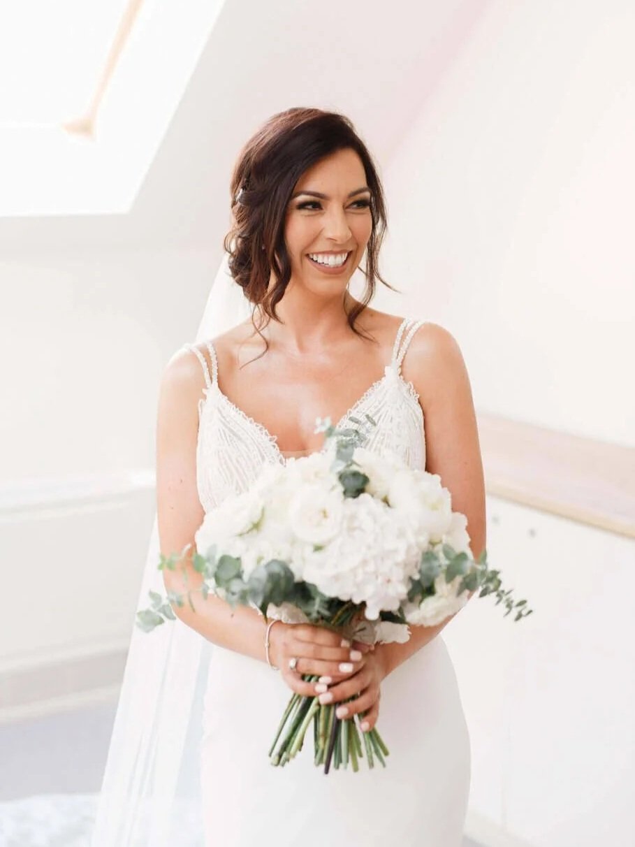 A woman in a wedding dress holding a bouquet of white flowers, smiling and looking off to the side.