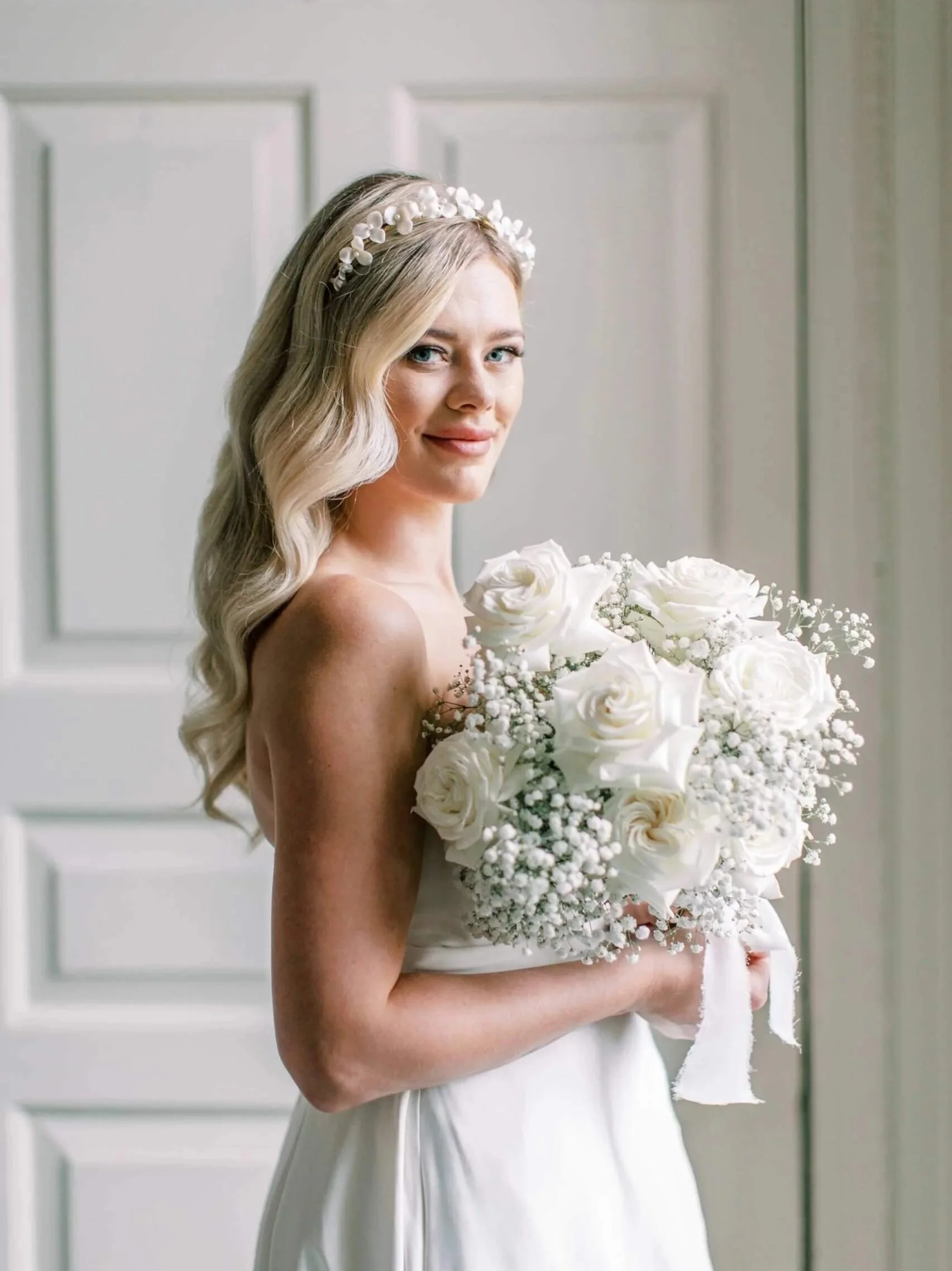 A young woman in a white wedding dress holding a bouquet of white flowers, standing in front of a paneled door.