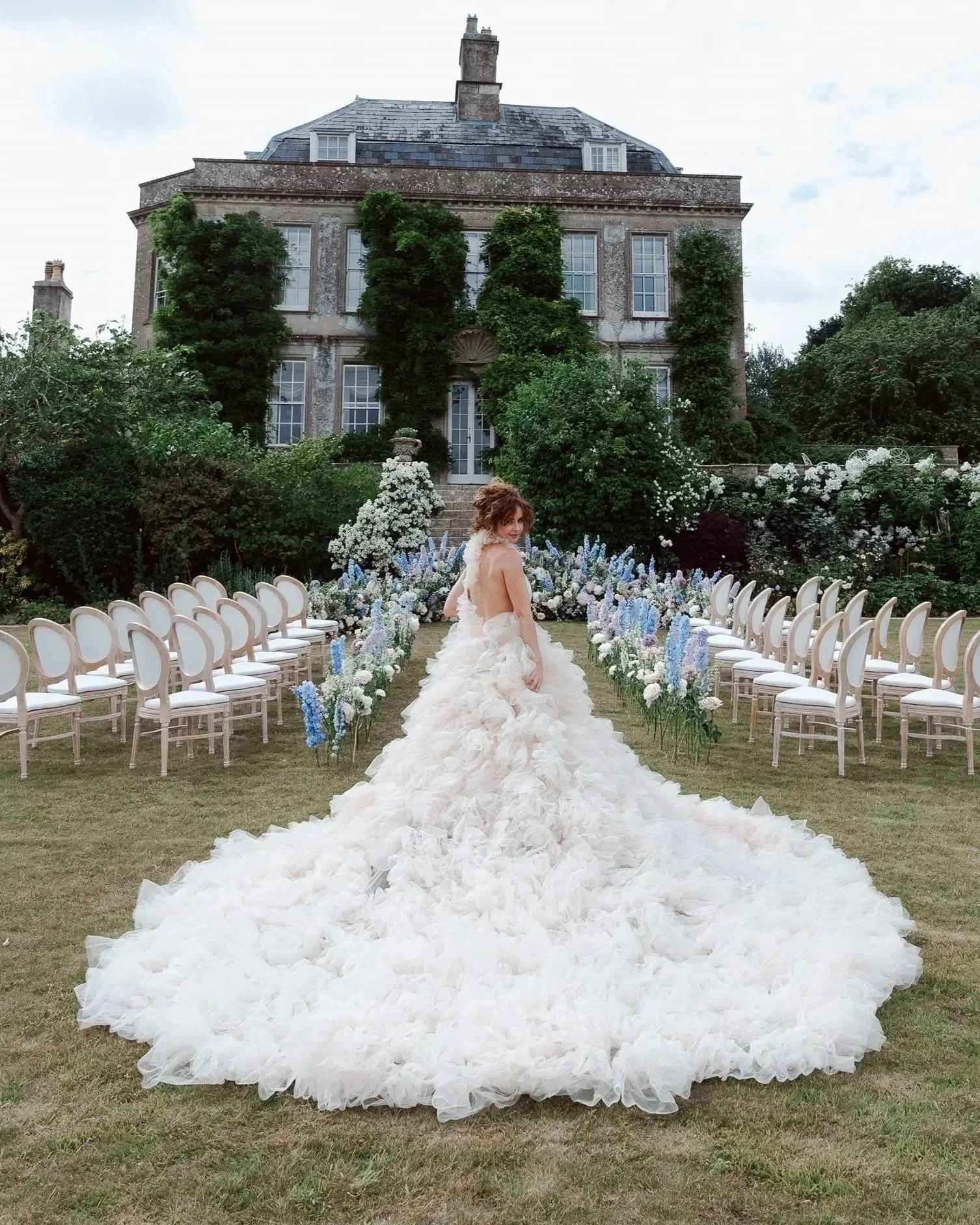 A woman in a white ruffled wedding gown with a long train standing in an outdoor garden wedding setup in front of a large, ivy-covered historic brick house with multiple windows and a chimney. The garden is decorated with white and blue flowers, and 