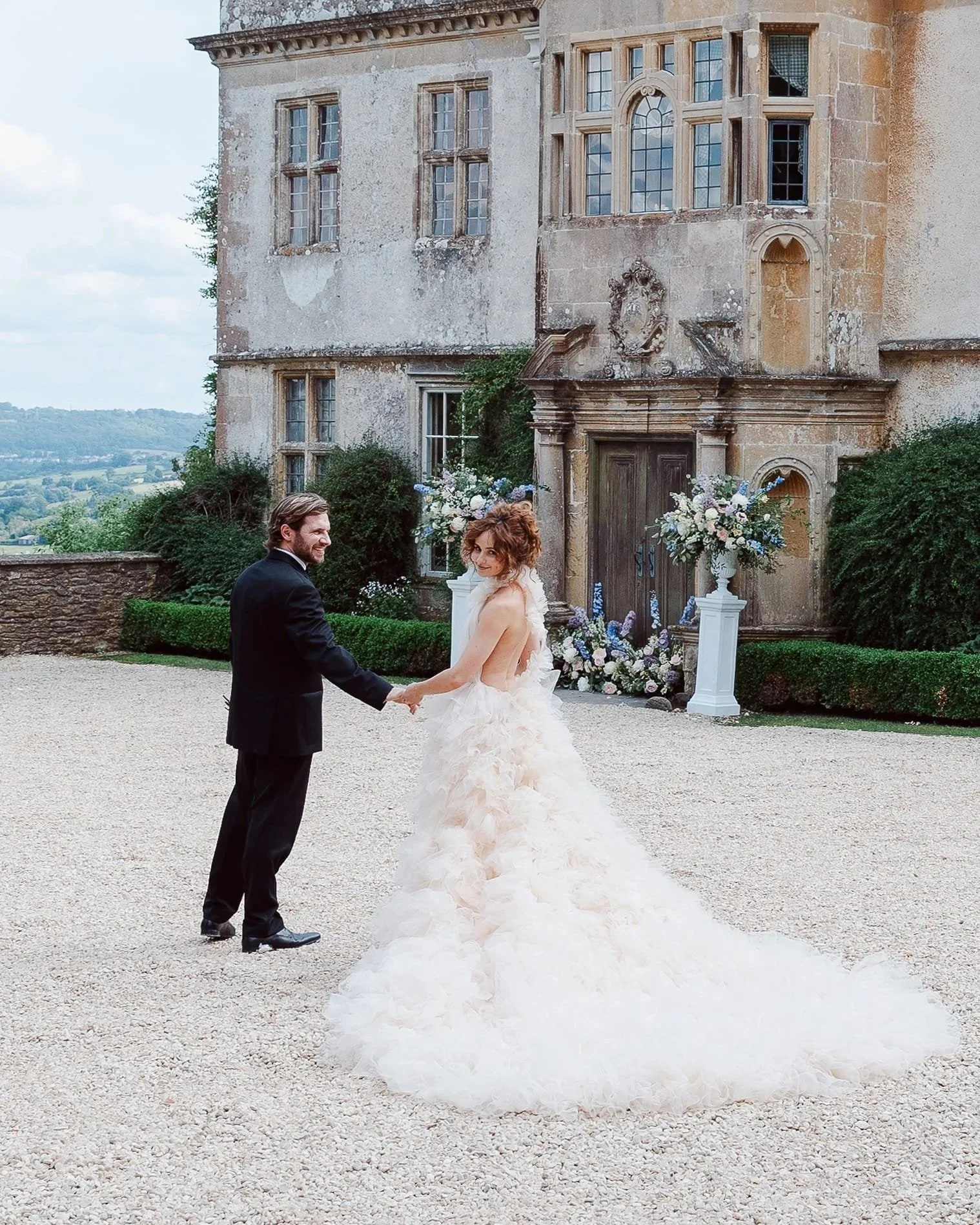 A bride and groom holding hands outside a historic stone building with large windows, flowers, and greenery.