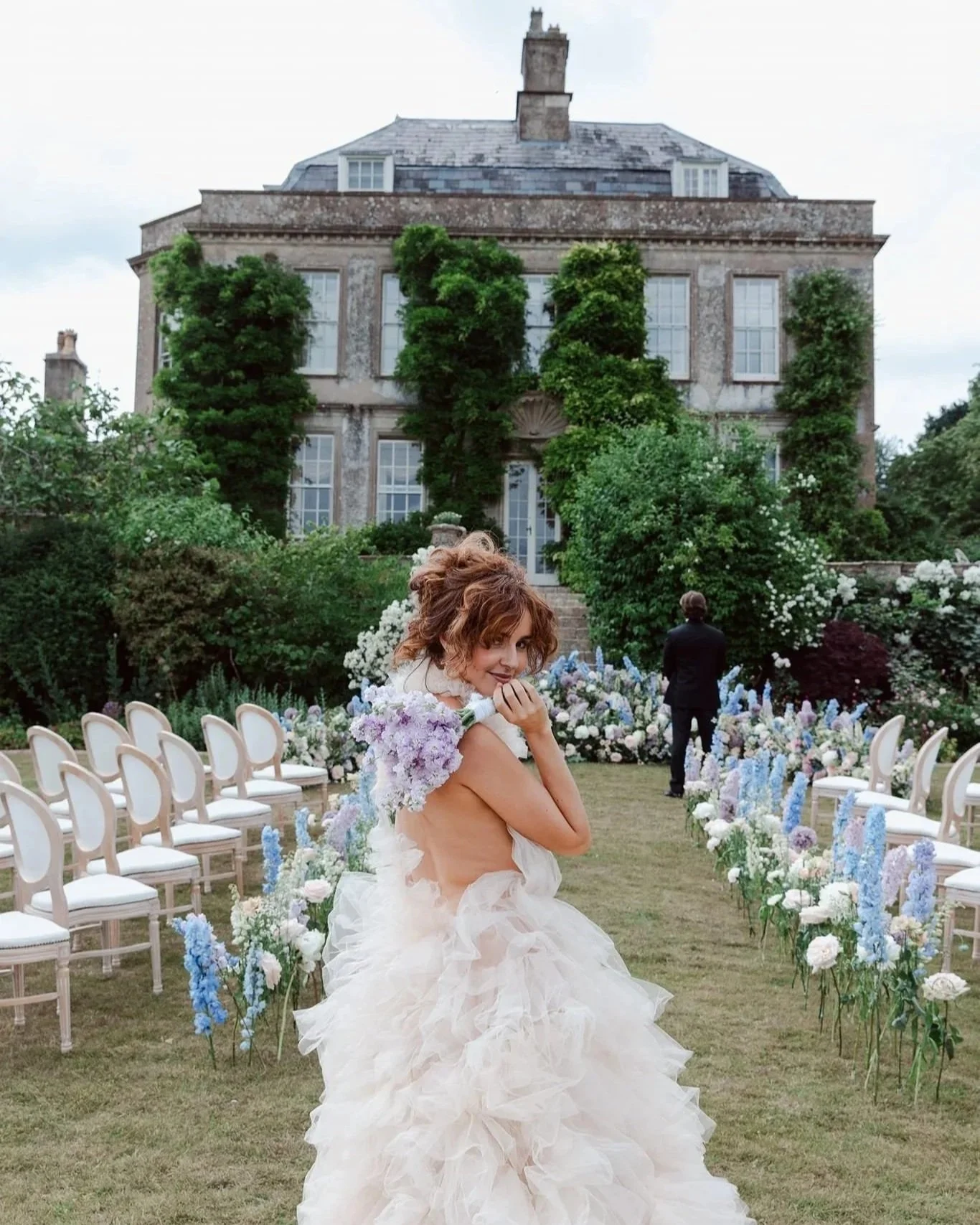 A woman in a ruffled white wedding dress holding purple flowers on her shoulder at an outdoor garden wedding, with rows of white chairs and an old stone mansion in the background.